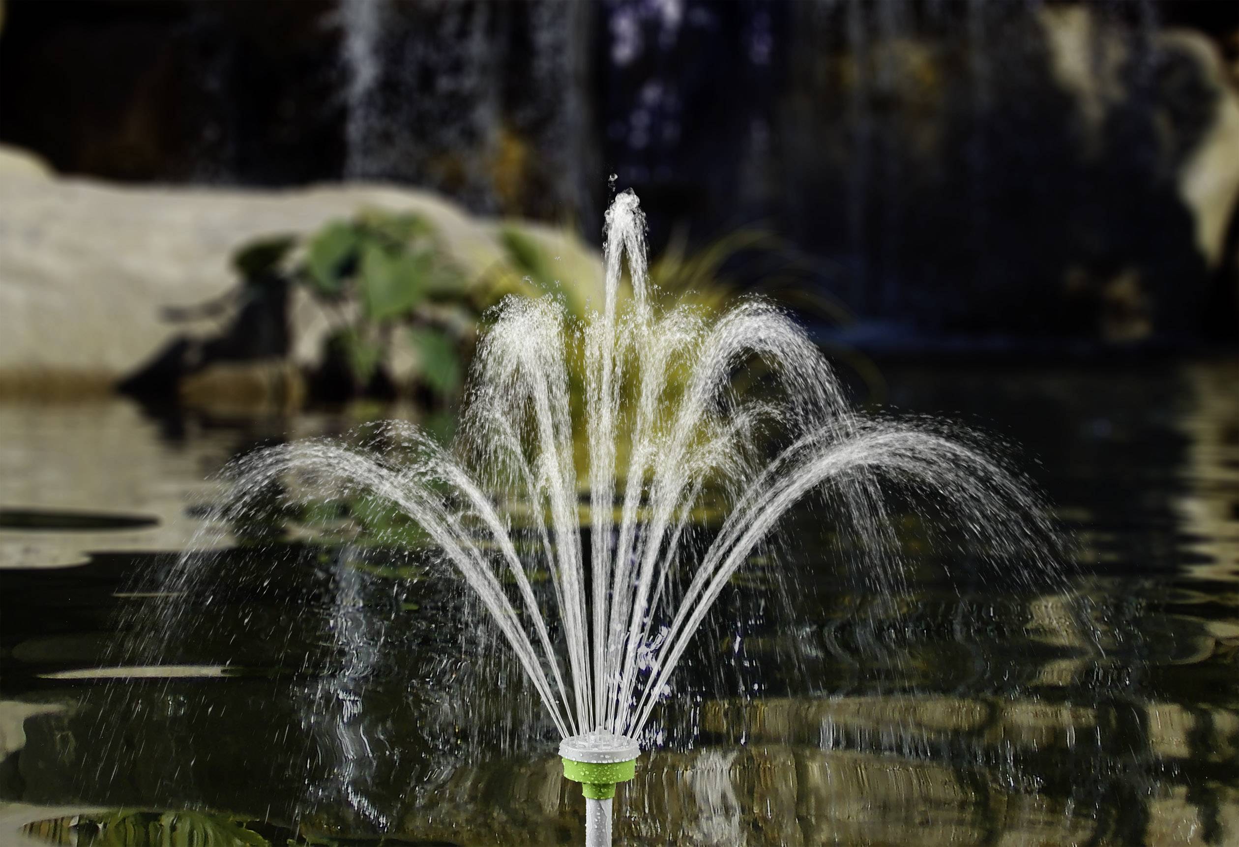 Fontaine avec des jets d'eau arqués dans un bassin ; des plantes et une cascade visibles en arrière-plan.