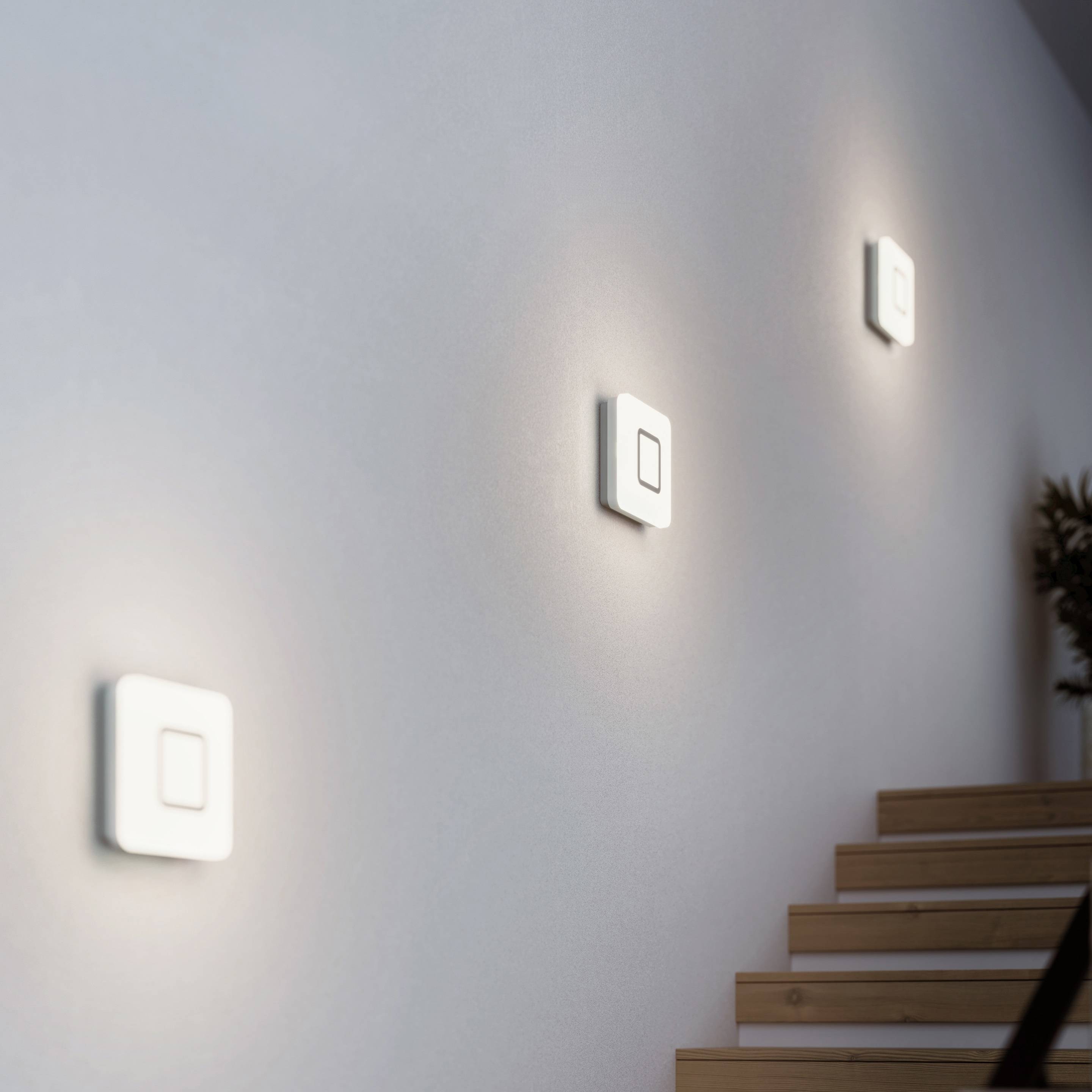 Trois appliques murales carrées éclairent un escalier de gauche à droite. Les luminaires sont répartis de manière uniforme et diffusent leur lumière vers l'extérieur.