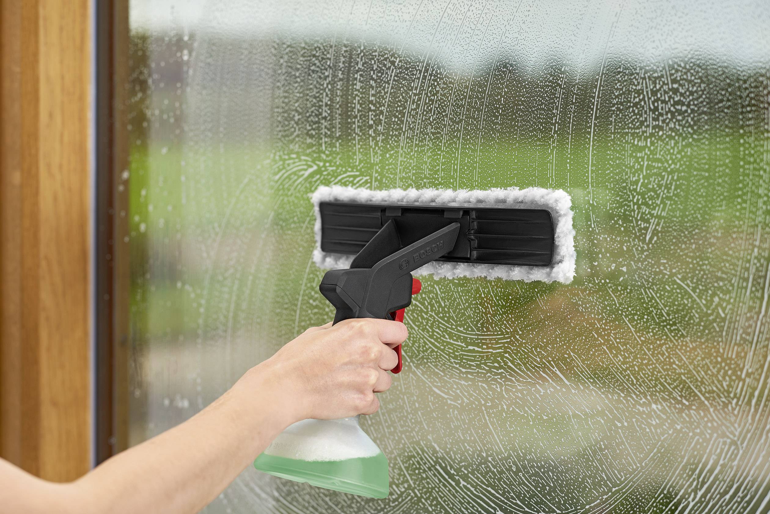 Une main nettoie une fenêtre avec un raclette et un spray nettoyant. À l'arrière-plan, un jardin vert flou est visible.