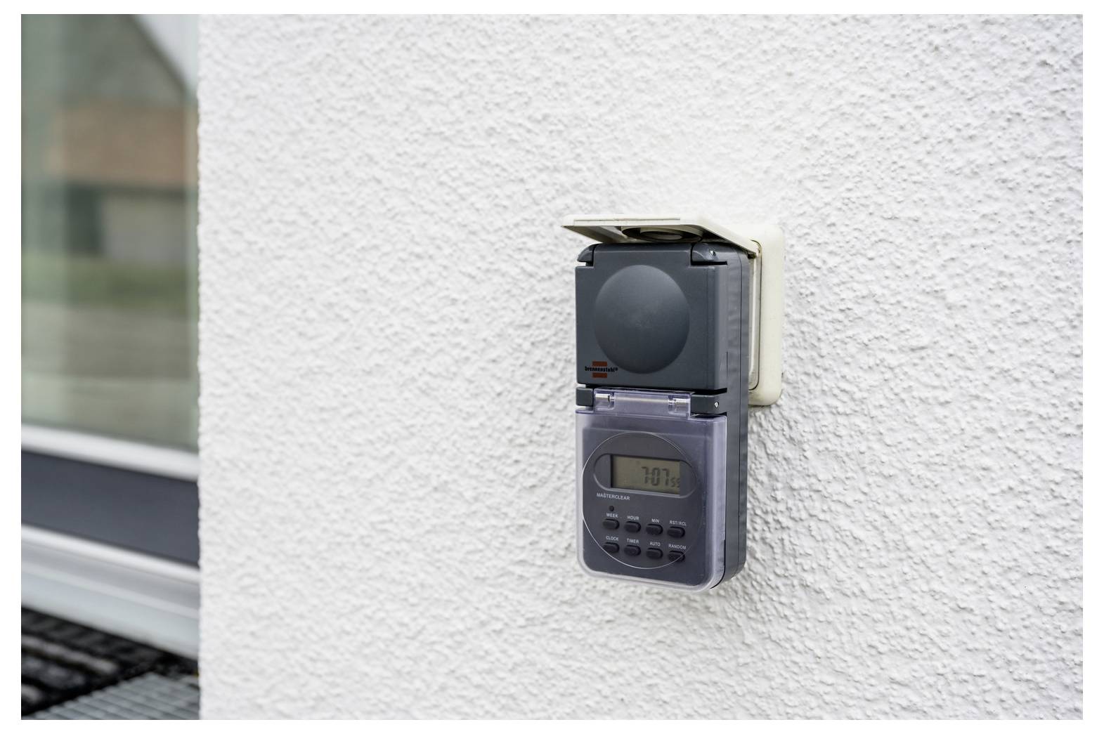 Minuteur numérique mural ou contrôleur de prise électrique sur un mur extérieur blanc à texture, avec un petit écran d'affichage et des boutons sous un couvercle de protection.