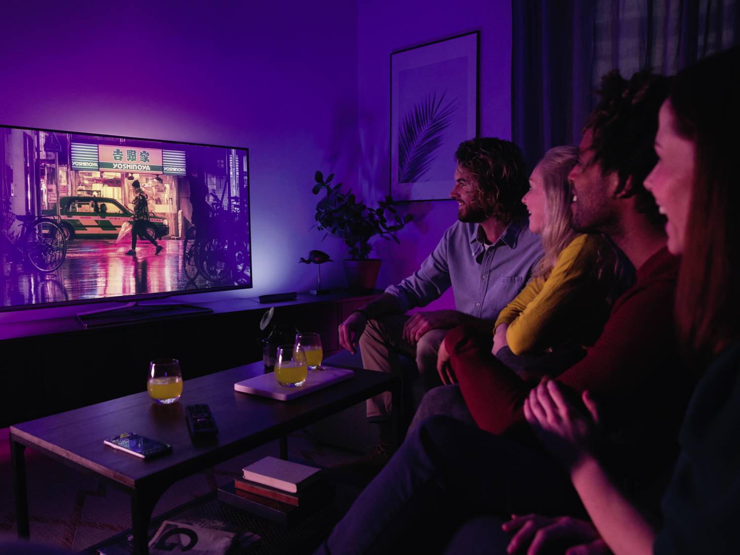 Un groupe de quatre personnes est assis sur un canapé et regarde ensemble un film sur un téléviseur dans une pièce obscurcie.