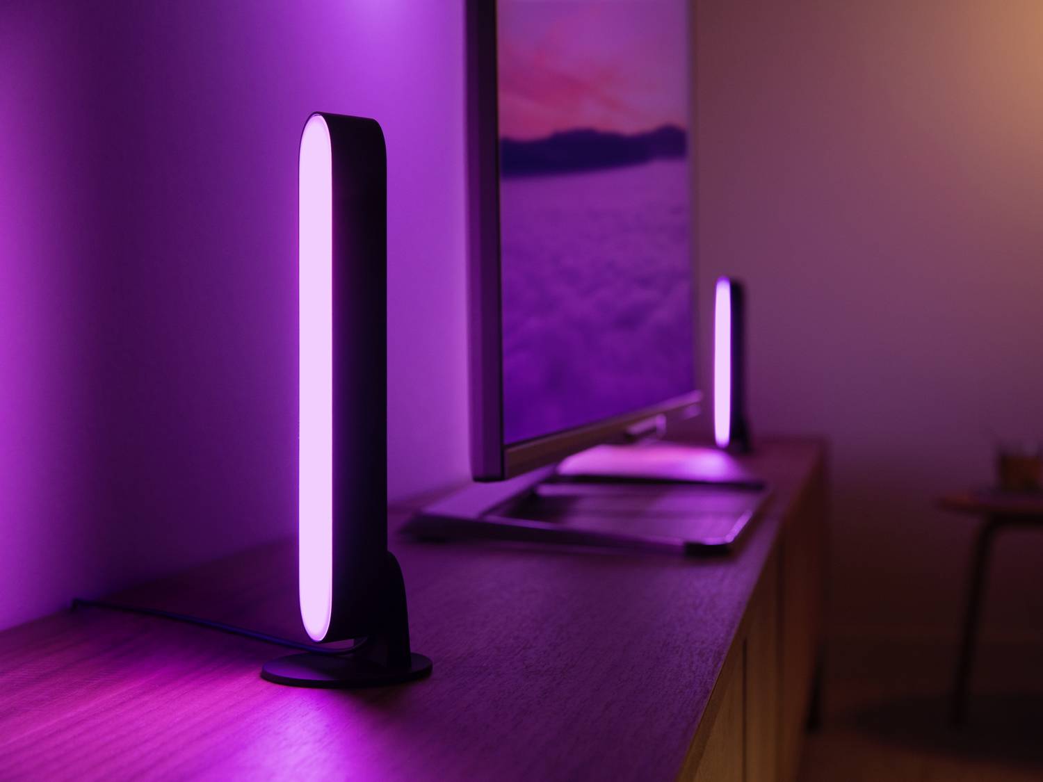 Deux lampes LED verticales émettant une lumière violette, placées de part et d'autre d'un écran sur un bureau dans une pièce obscure.