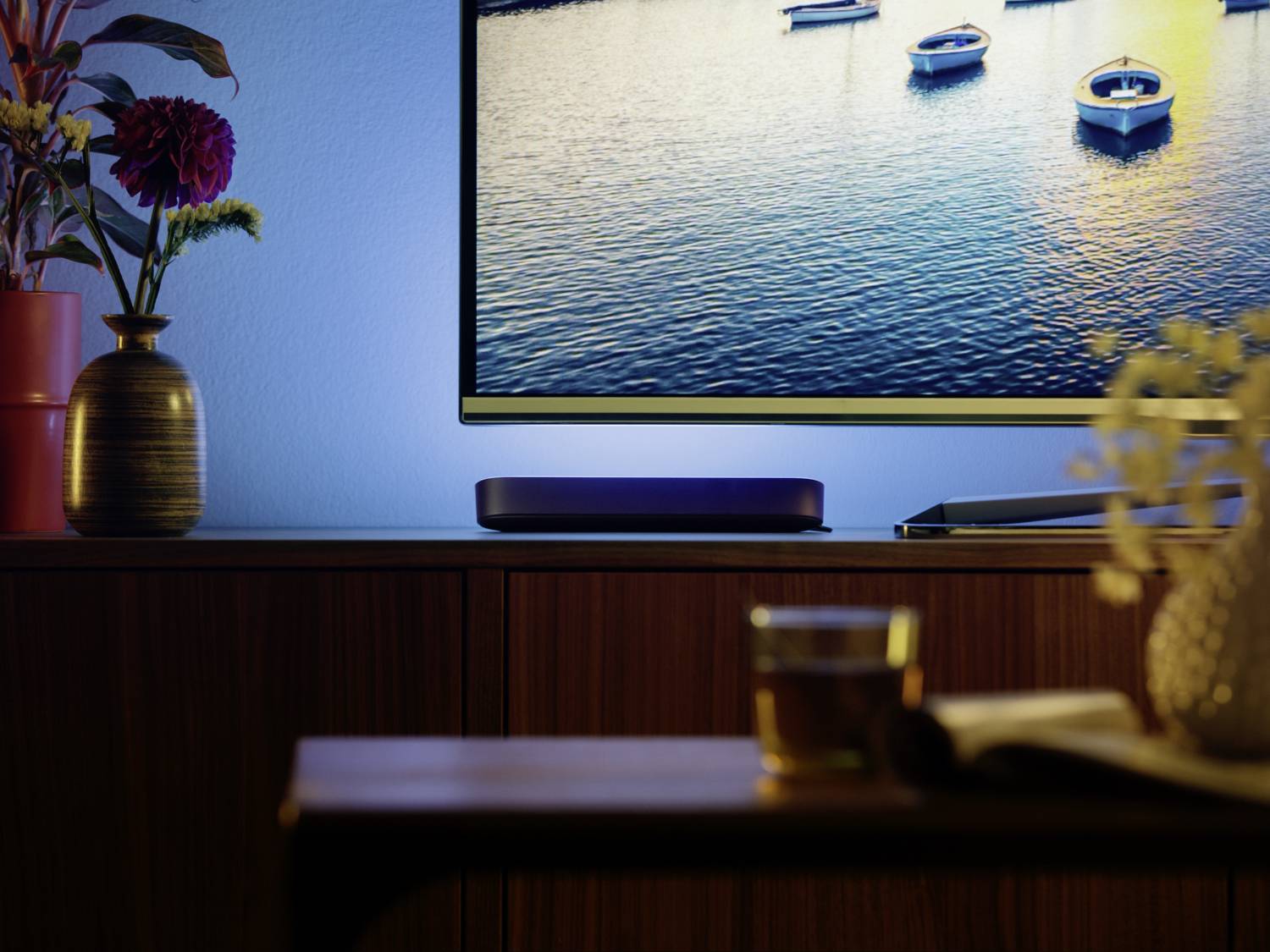 Au premier plan, un verre de thé et un livre sur une table. En arrière-plan, un téléviseur affichant l'image d'un lac, avec des fleurs sur une commode.