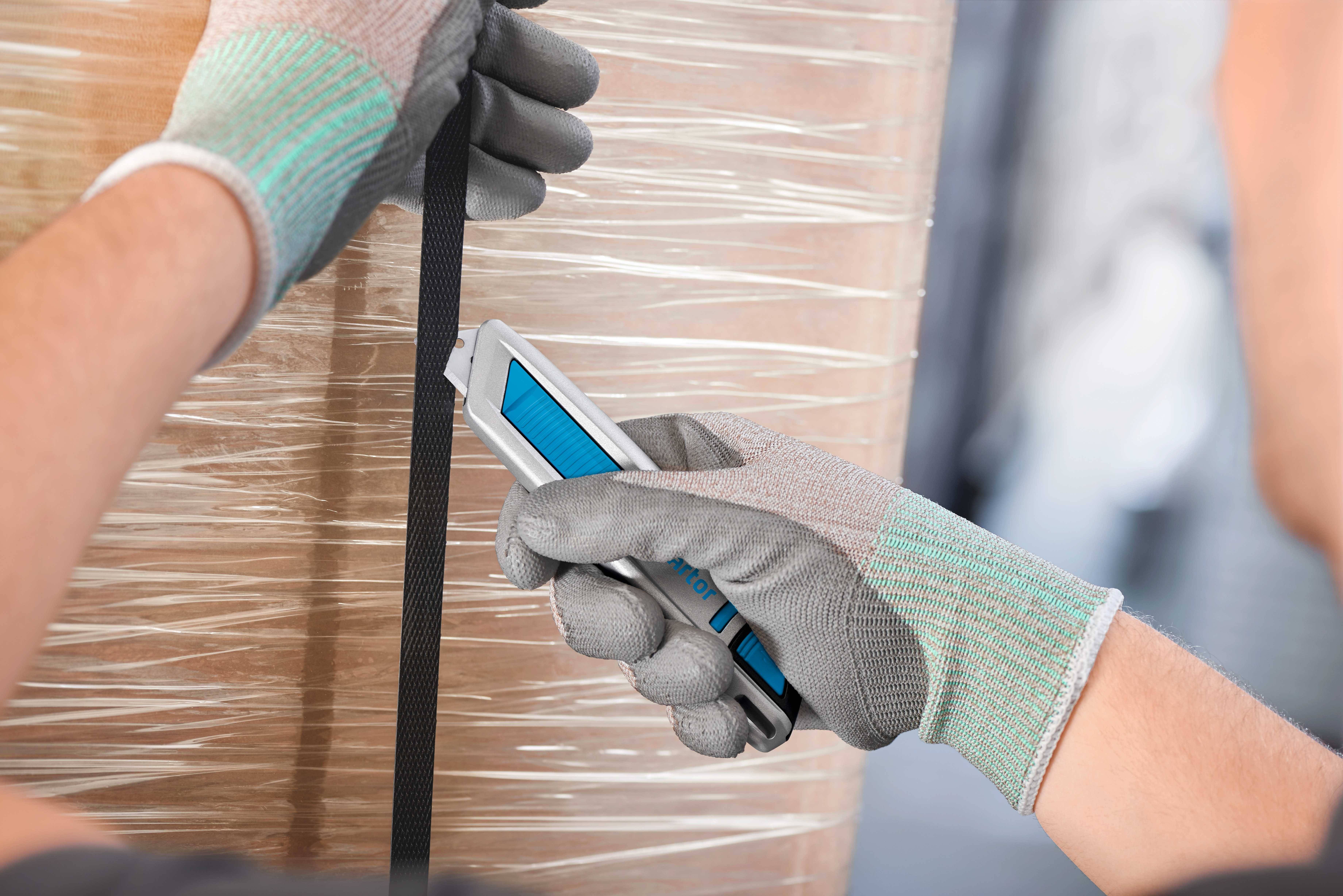 Une personne portant des gants gris coupe avec un cutter bleu un ruban noir autour d'un emballage.