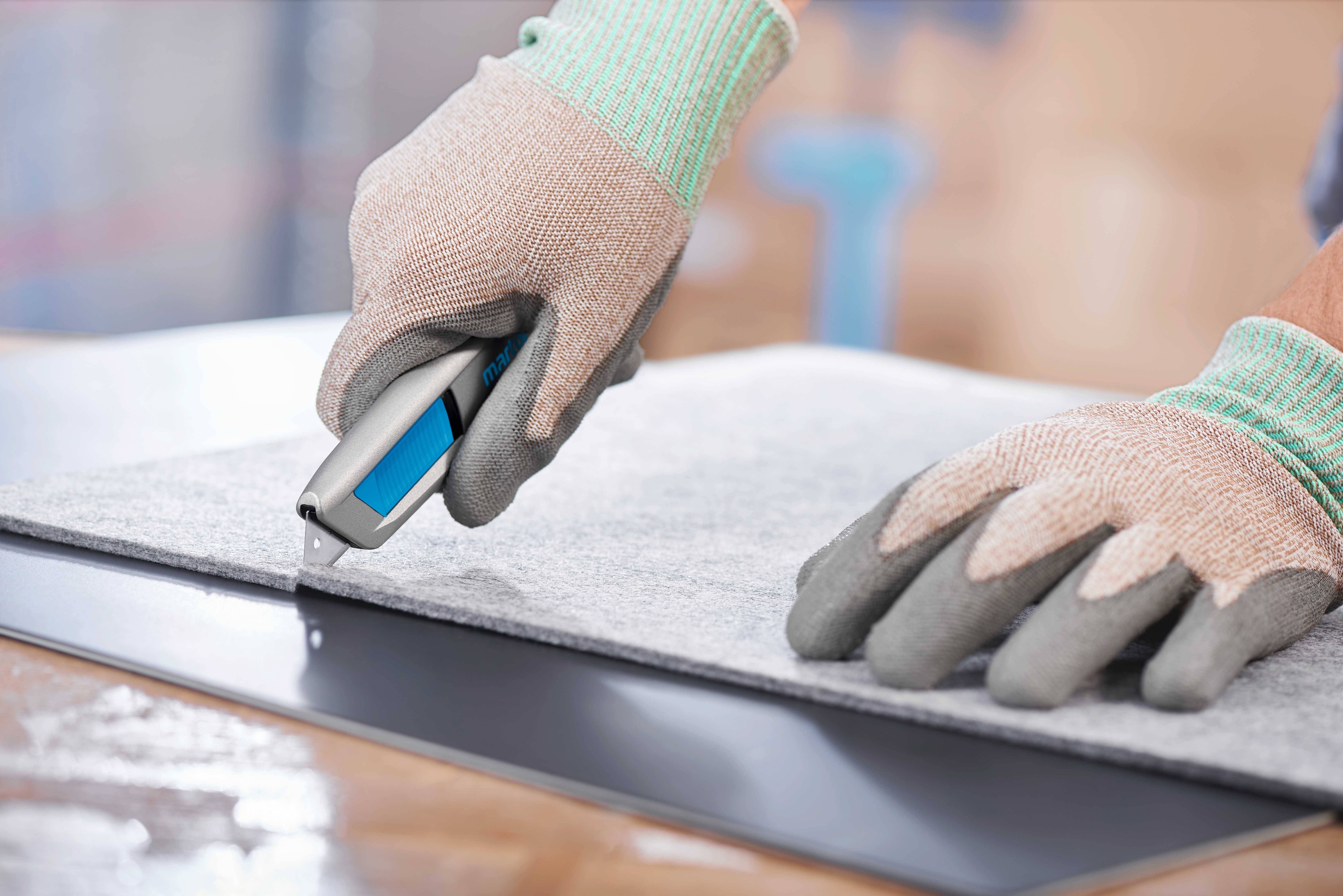 Une personne coupe une natte grise avec un couteau à tapis, portant des gants de sécurité.