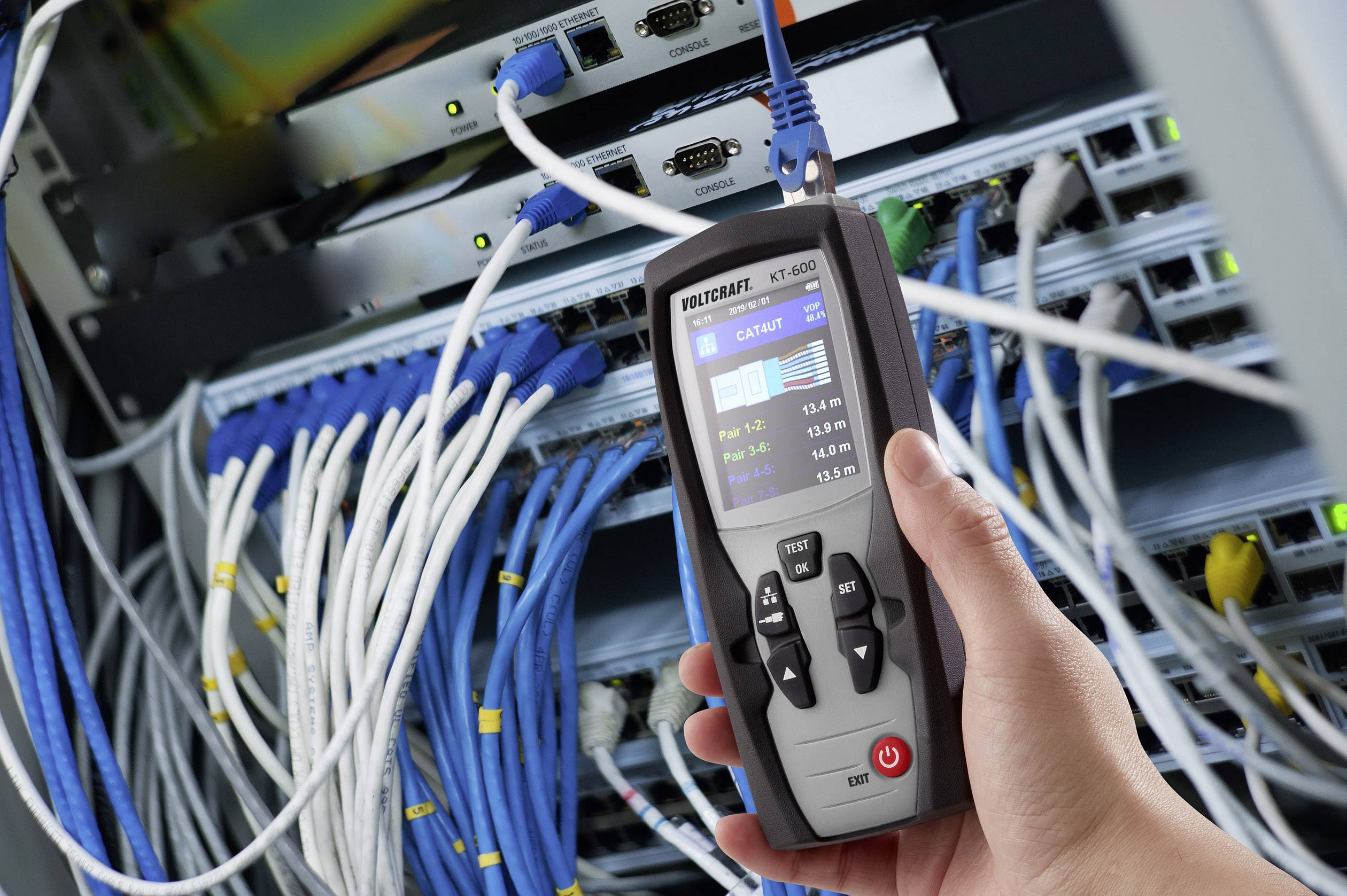 Une main tient un appareil d'analyse réseau devant une armoire de serveurs remplie d'équipements réseau câblés, afin de vérifier les connexions.