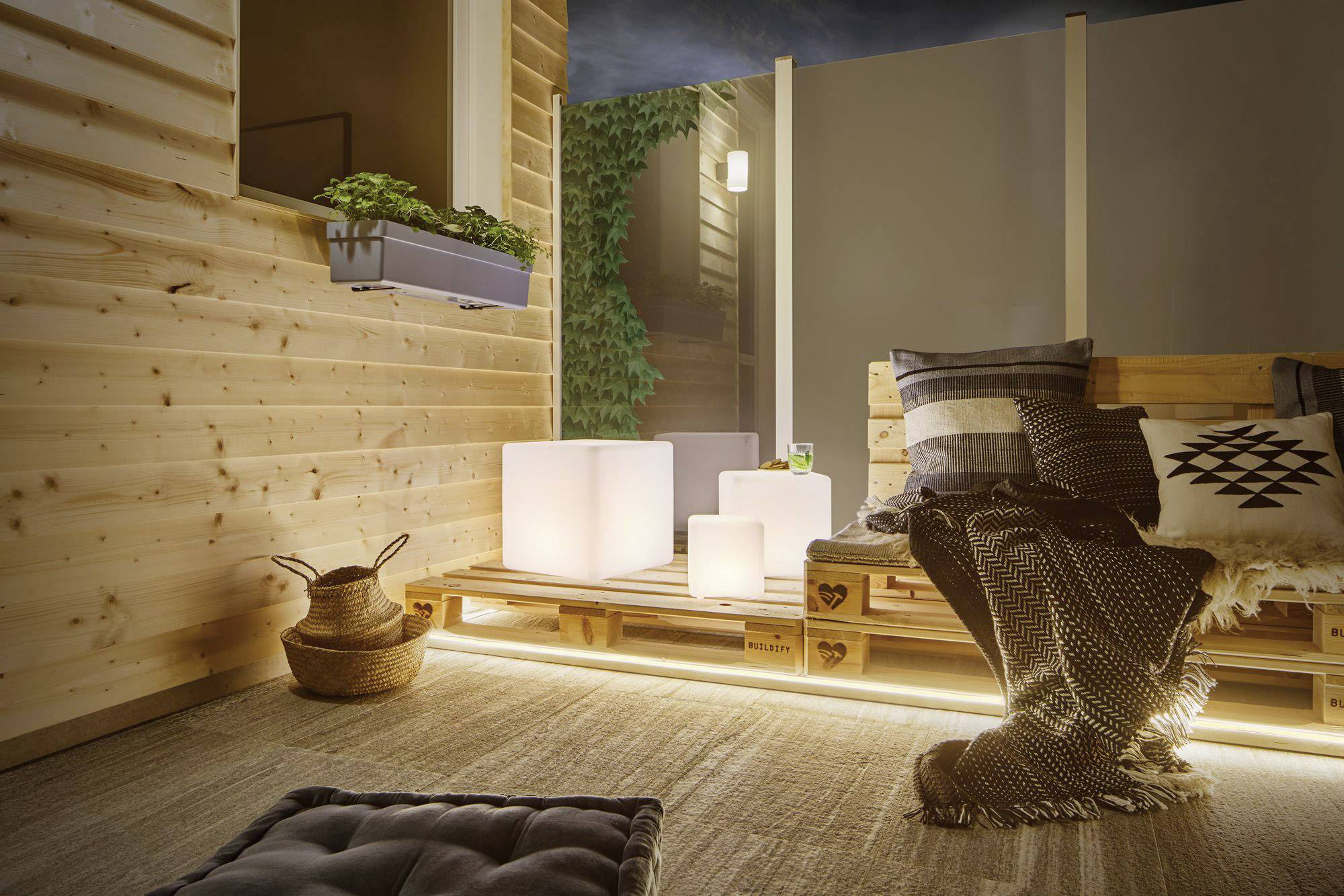 Terrasse chaleureuse avec des murs et des meubles en bois. Coussins décoratifs et couvertures, éclairage doux par des cubes LED et présence de plantes.