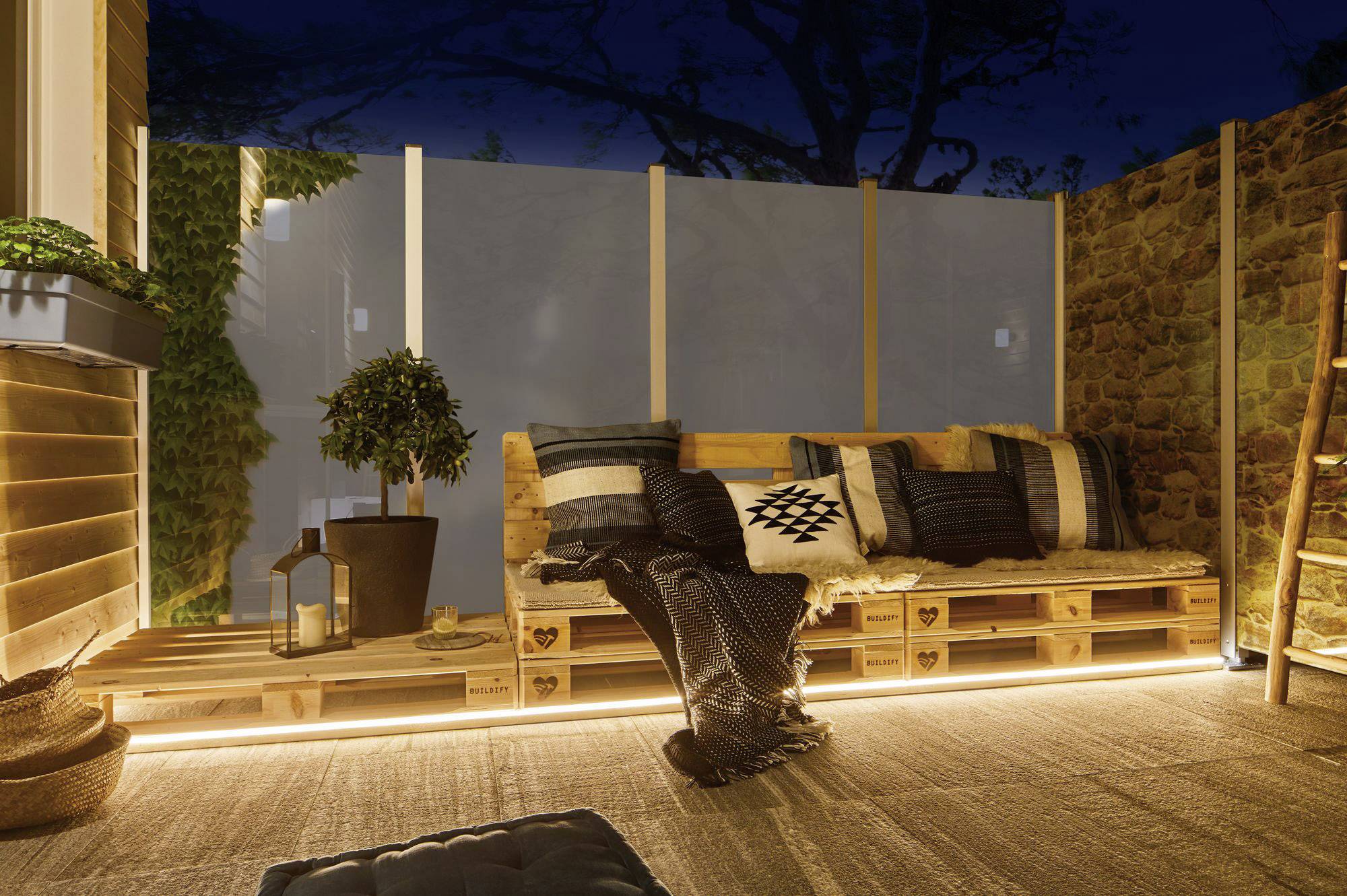 Terrasse accueillante de nuit avec des meubles en bois, des coussins et un éclairage. Une lanterne et des plantes créent une atmosphère chaleureuse et invitante.