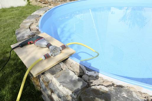 Une piscine est remplie à l'aide d'un tuyau connecté à une pompe. La pompe est installée sur une planche en bois au bord de la piscine.