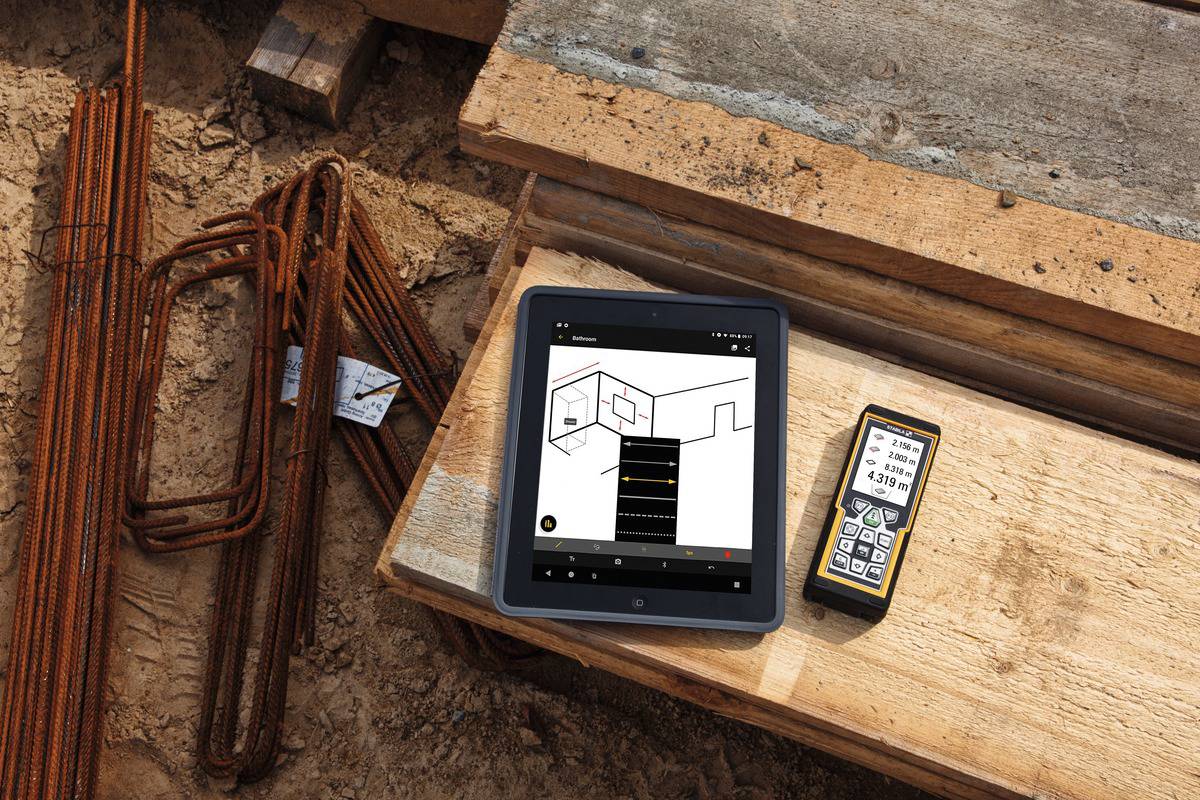 Une tablette et un appareil de mesure numérique sont posés sur un chantier de construction. La tablette affiche un plan de construction, l'appareil de mesure indique une mesure.