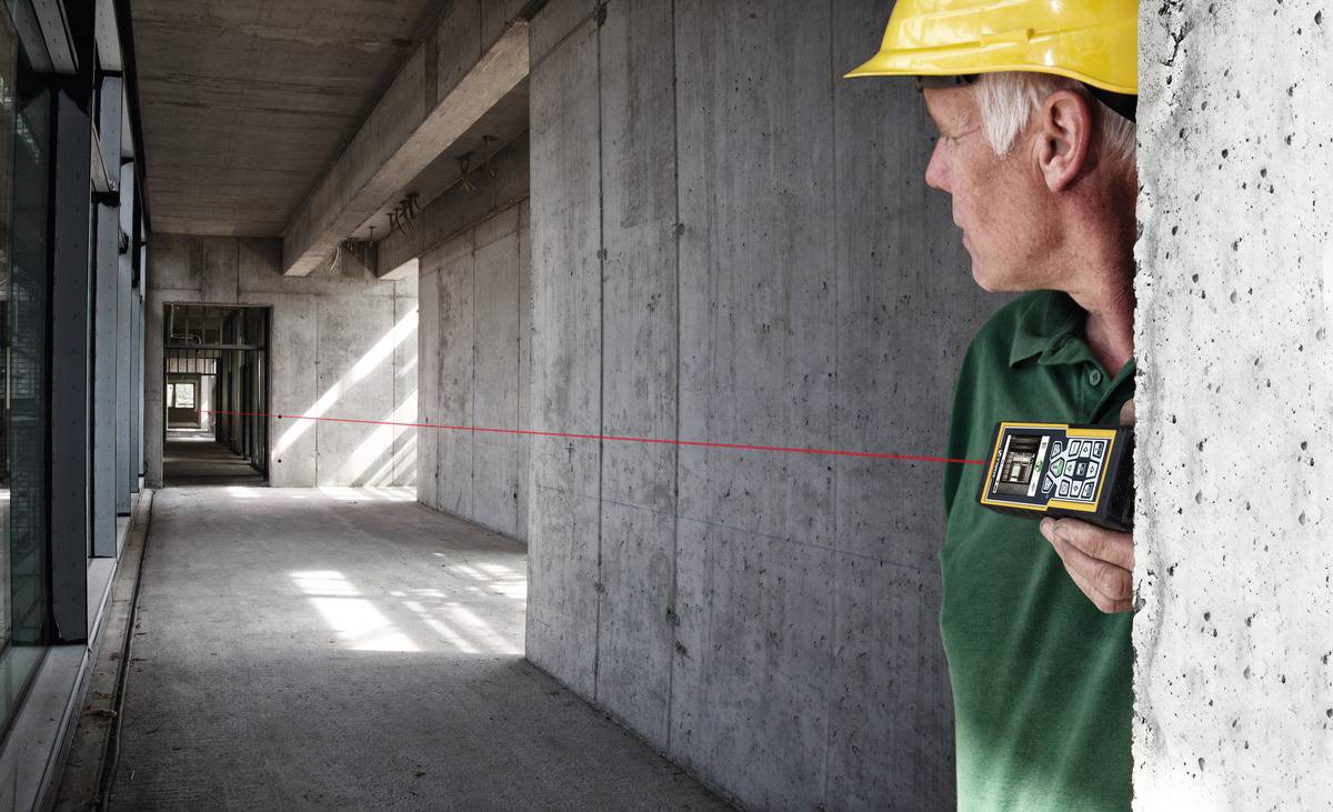 Un ouvrier portant un casque de chantier jaune mesure une longue halle en béton à l'aide d'un télémètre laser.