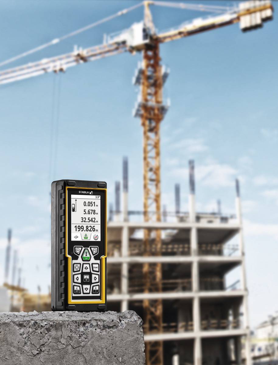 Un télémètre laser est posé sur une surface en béton, avec un bâtiment en construction et une grue en arrière-plan.