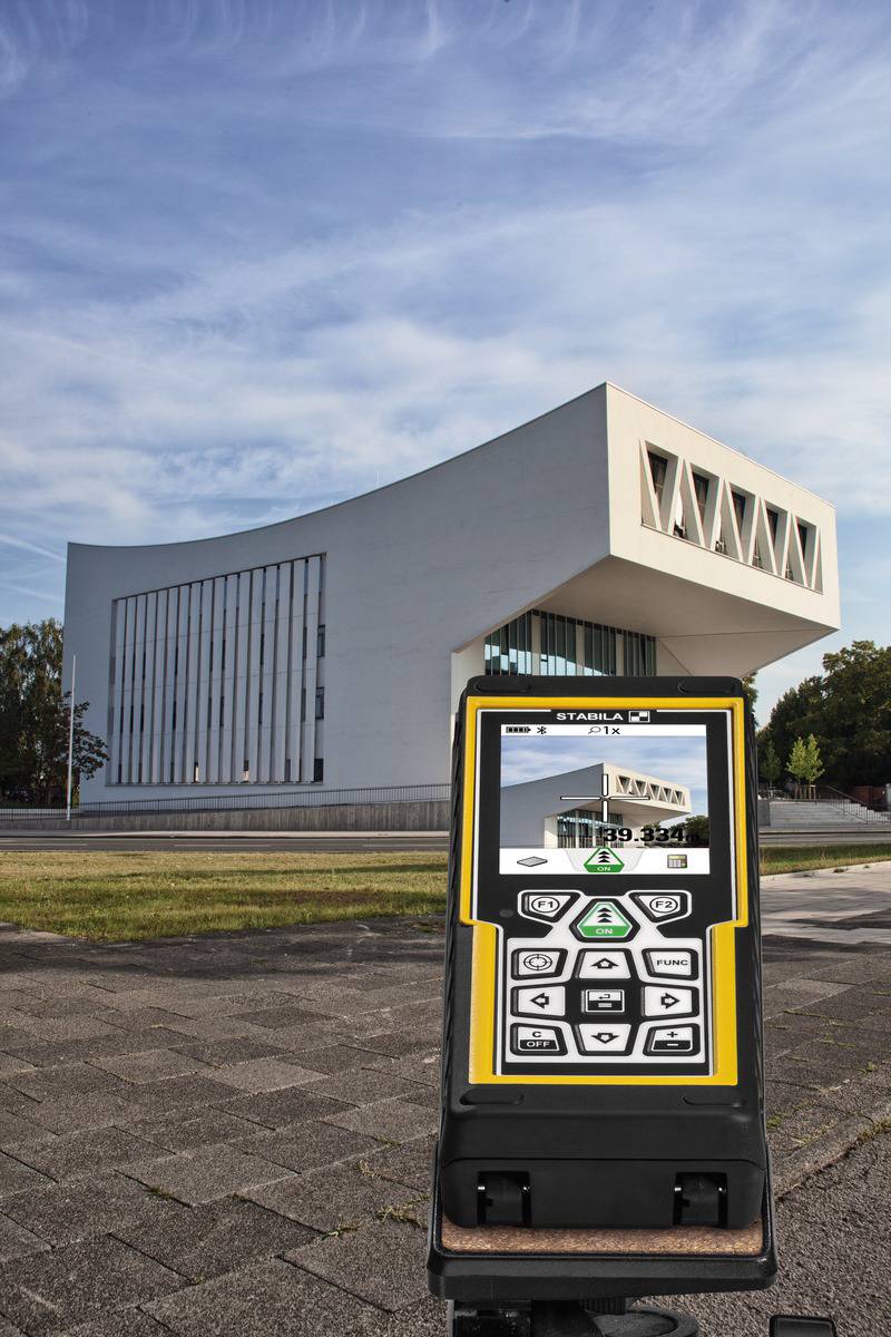 Un bâtiment blanc et moderne au toit incliné se dresse en arrière-plan, tandis qu'un télémètre laser est visible au premier plan.