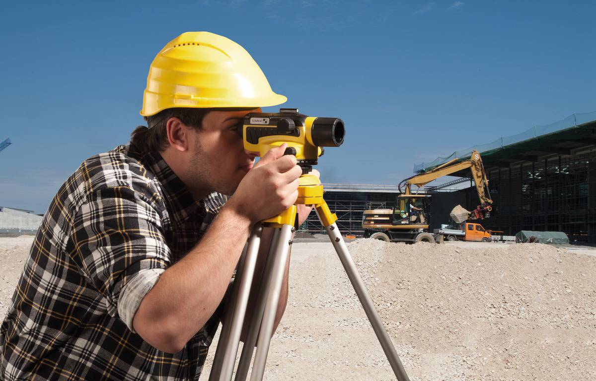 Un ouvrier portant un casque de chantier jaune utilise un instrument de mesure sur un chantier. Un excavateur est visible en arrière-plan.