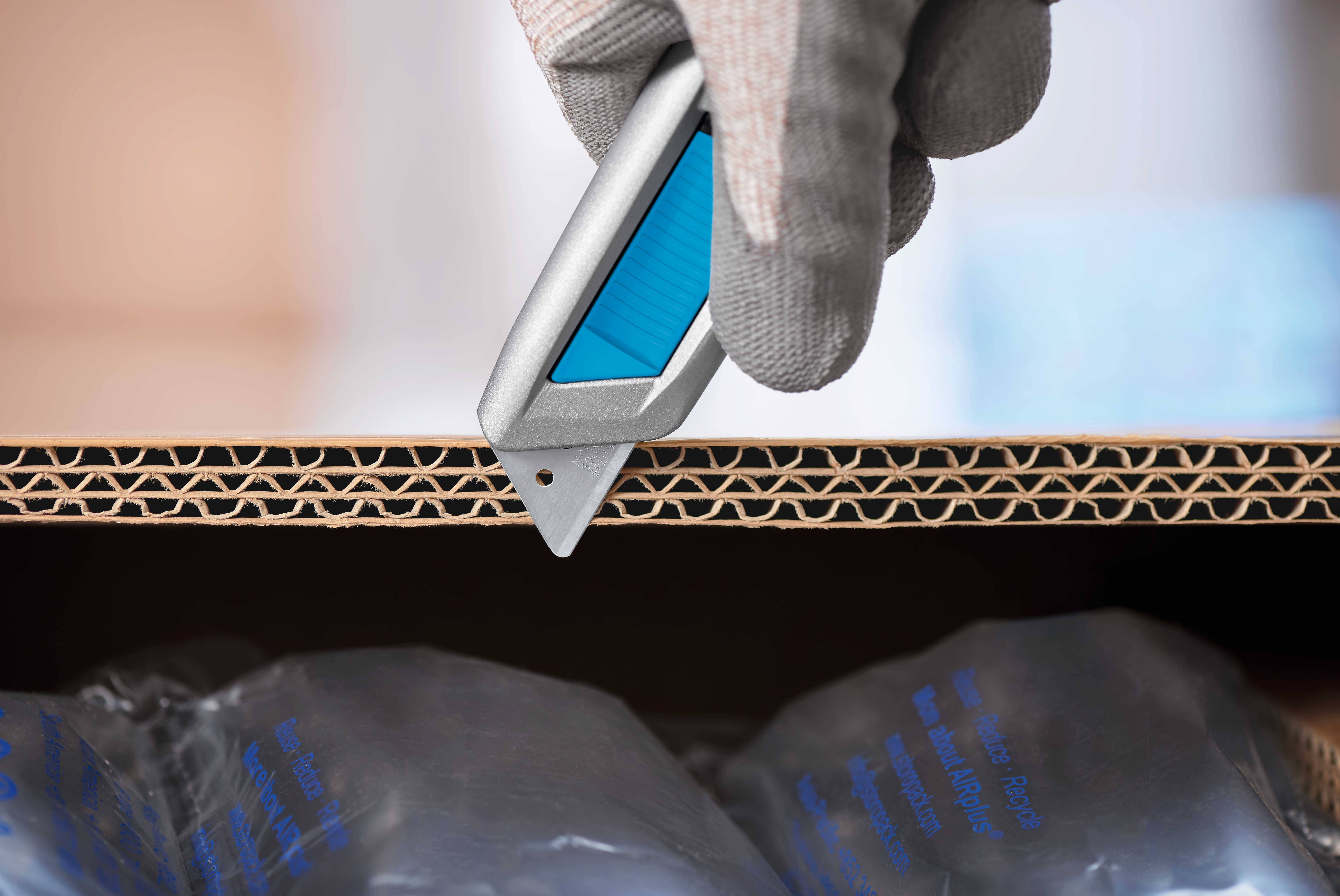 Une main gantée coupe un carton avec un couteau. Des emballages en plastique sont visibles en dessous.