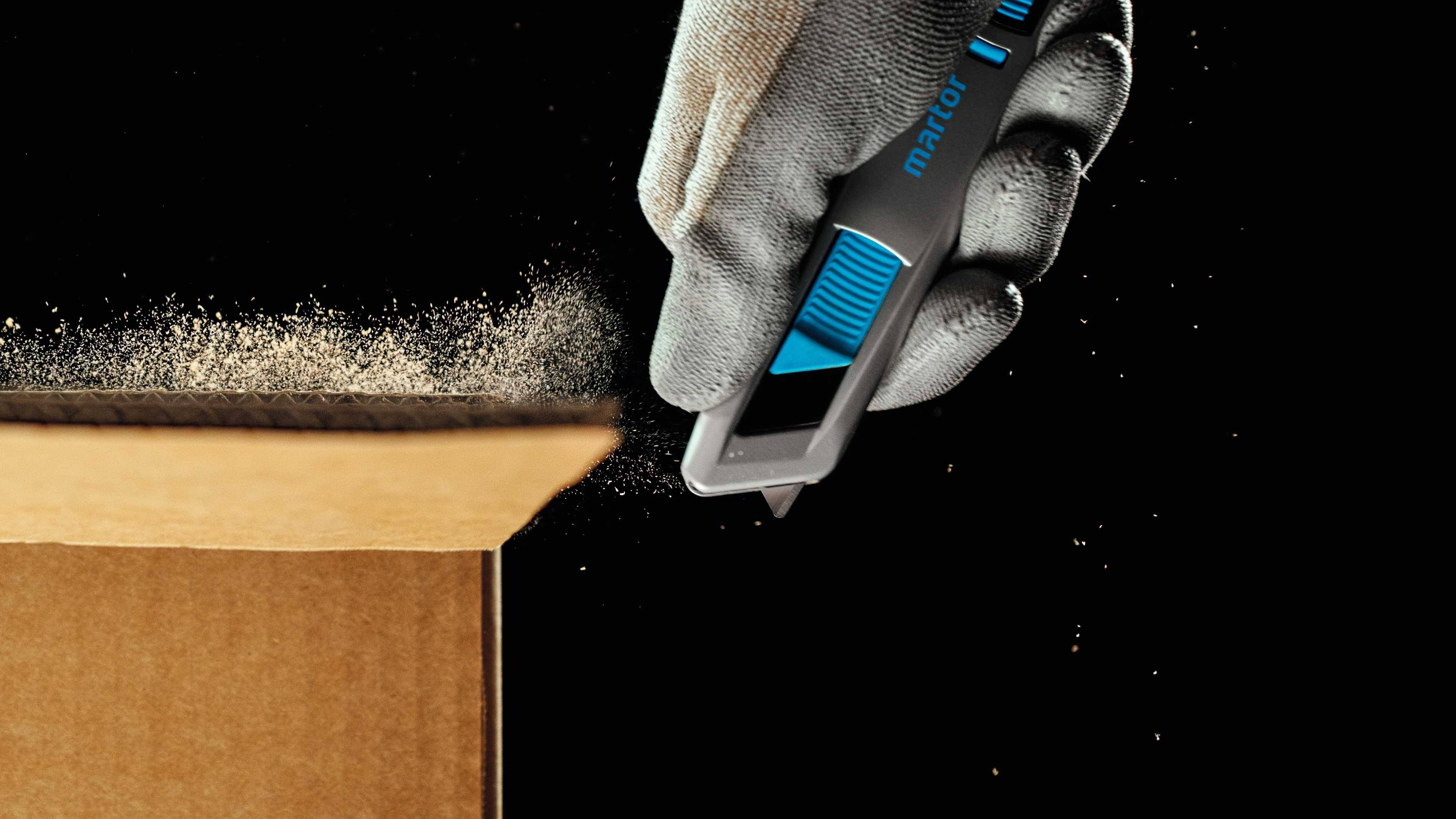 Une main gantée utilise un outil tranchant pour couper le bord d'un carton. Des particules de poussière flottent dans l'air.