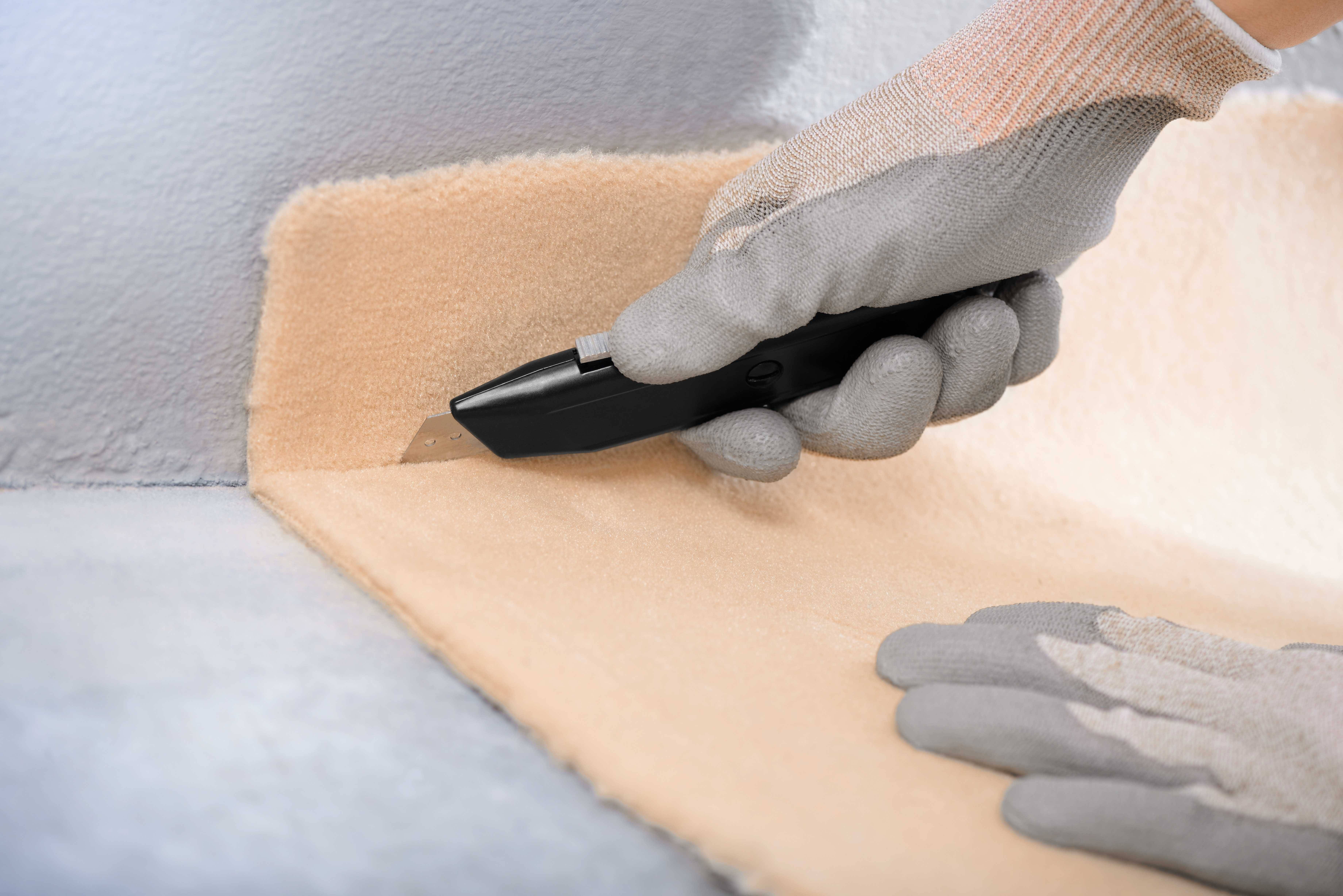 Une main gantée coupe un tapis beige le long d'un bord avec un cutter noir.