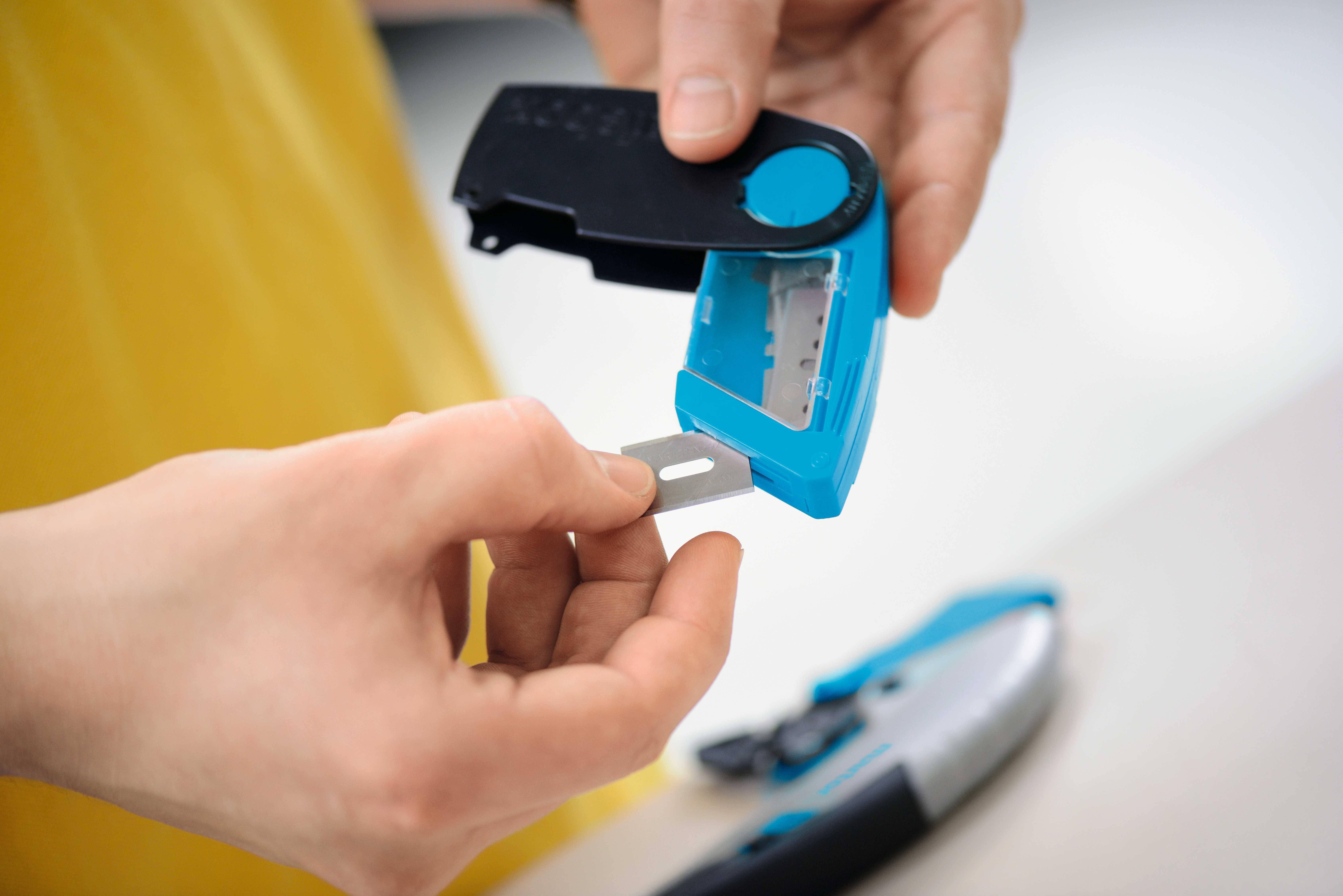 Une personne ouvre un boîtier pour insérer une lame de rechange dans un cutter bleu. Les mains tiennent la lame et le boîtier.