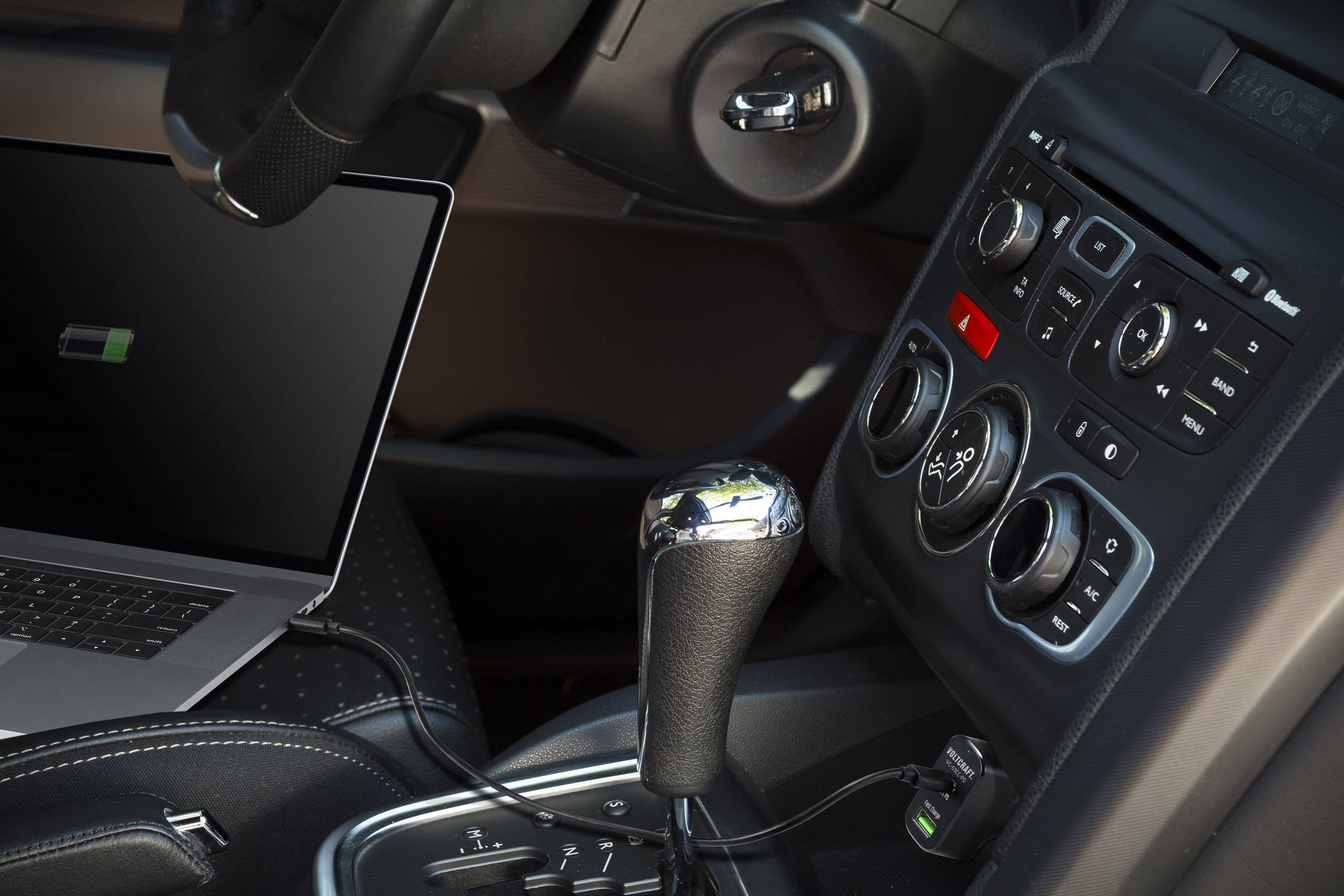 Un ordinateur portable se charge dans la voiture, branché au tableau de bord. On peut voir le volant, la console centrale et le levier de vitesses.
