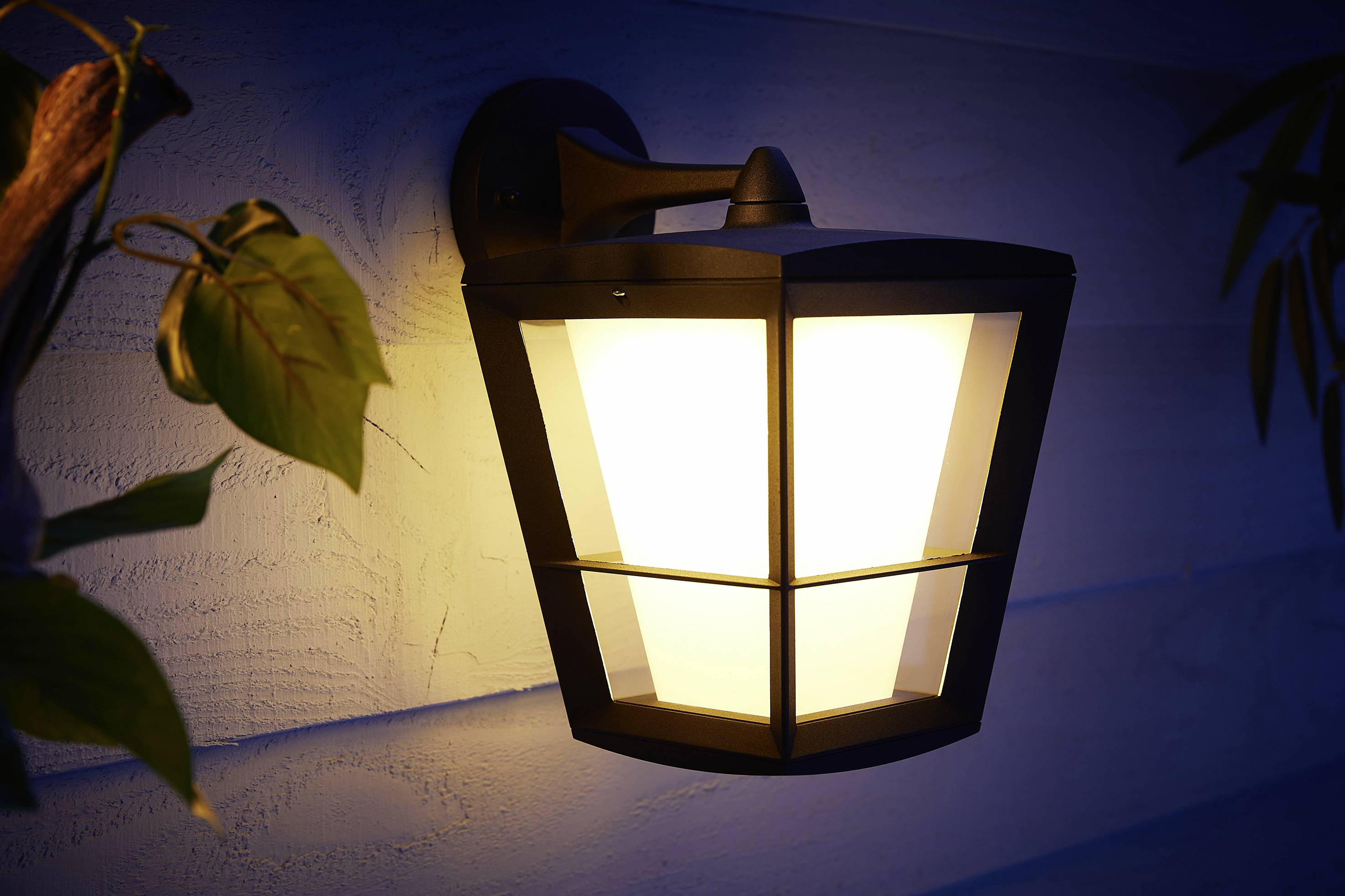 Lampe murale extérieure qui brille dans l'obscurité, installée sur une façade en bois avec quelques feuilles au premier plan.