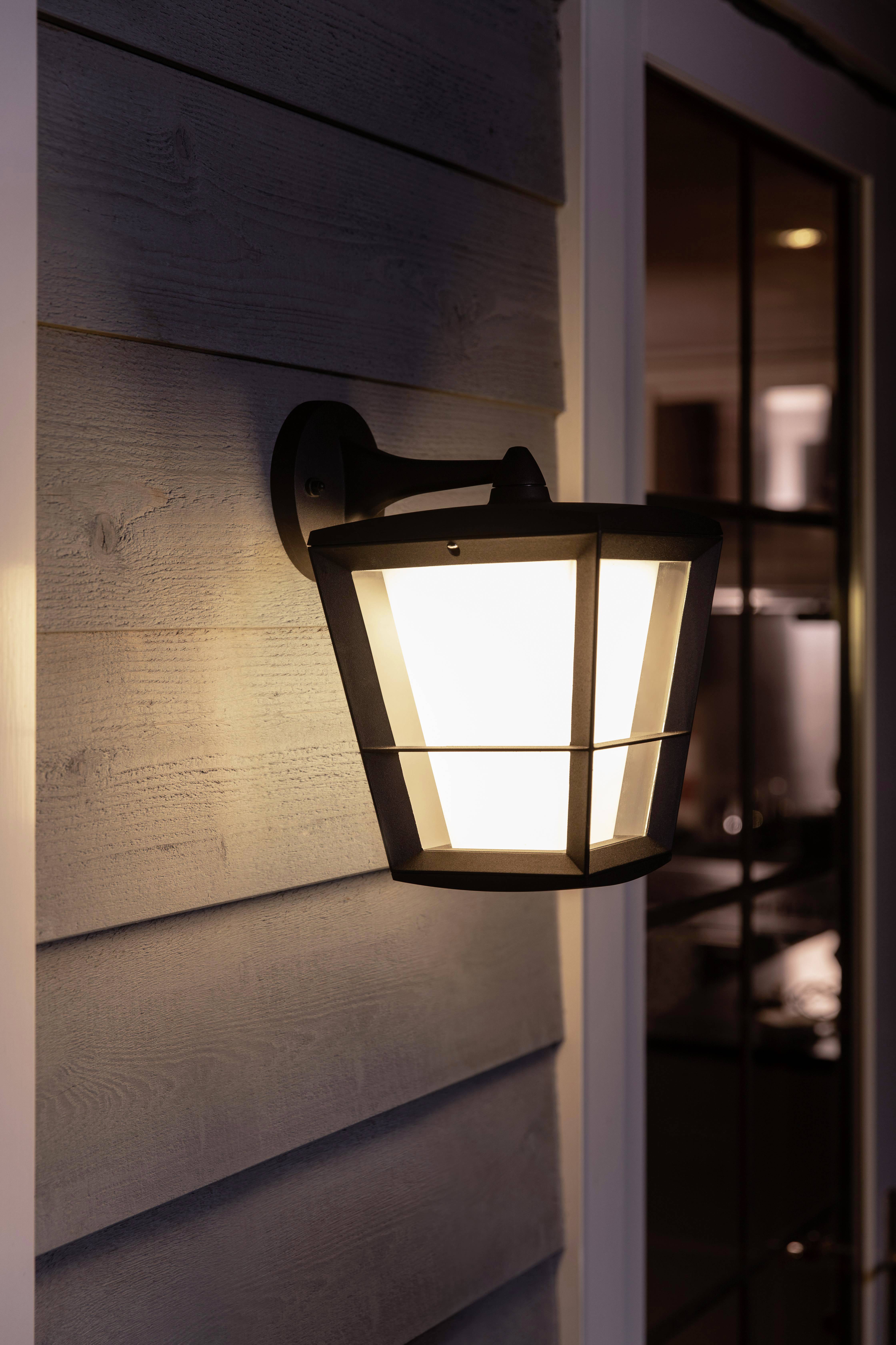 Lampe extérieure éclairant une véranda au crépuscule, fixée à un mur recouvert de lambris, à côté d'une porte vitrée.