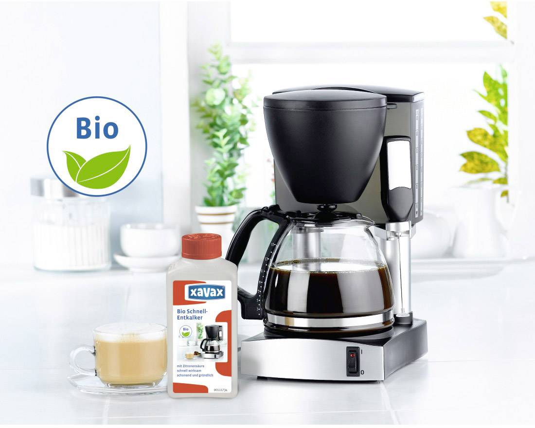 Une machine à café avec une carafe en verre pleine, à côté d'un paquet de détartrant bio et une tasse de café au lait. Arrière-plan : cuisine lumineuse.