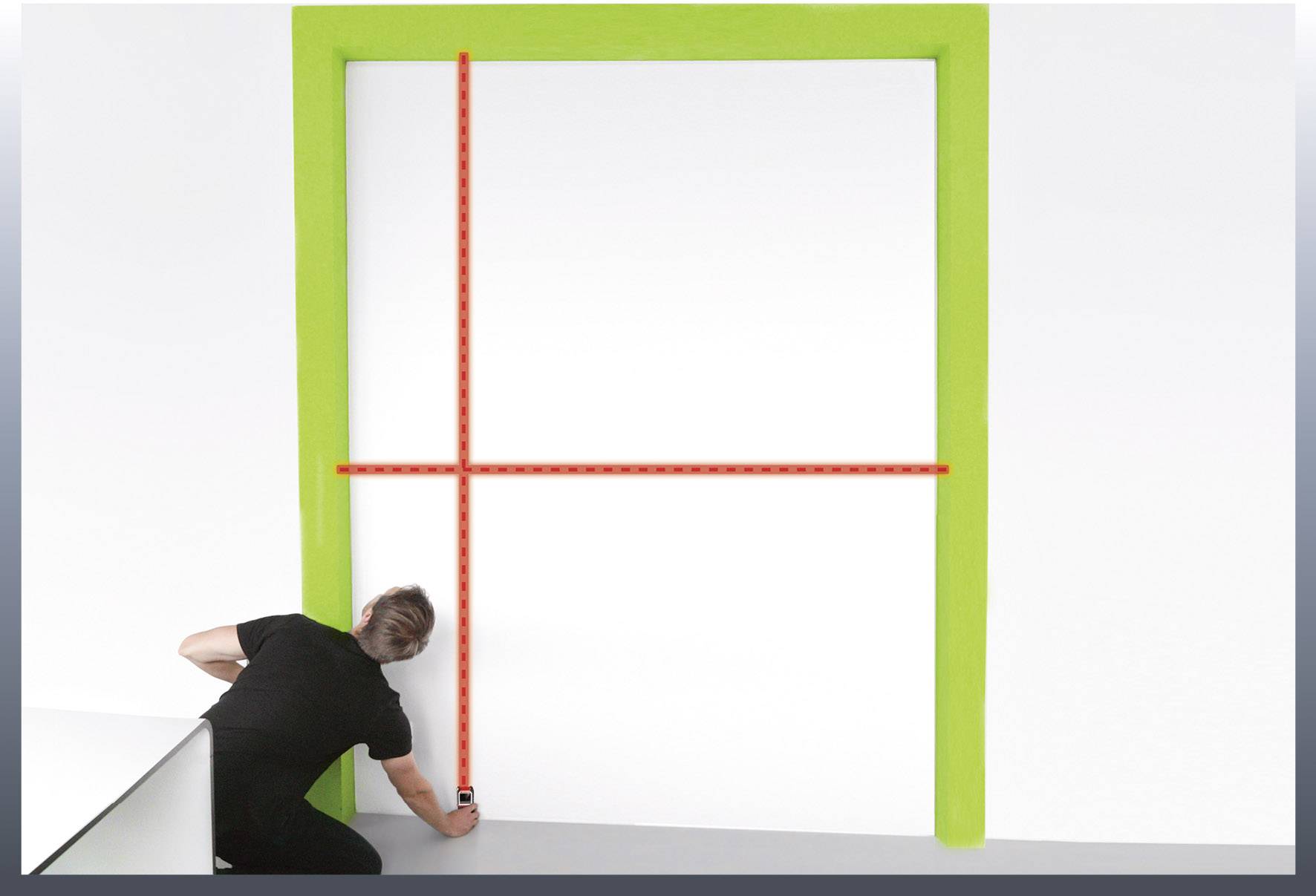 Un homme mesure l'intérieur d'un cadre rectangulaire vert avec un outil laser contre un mur blanc.