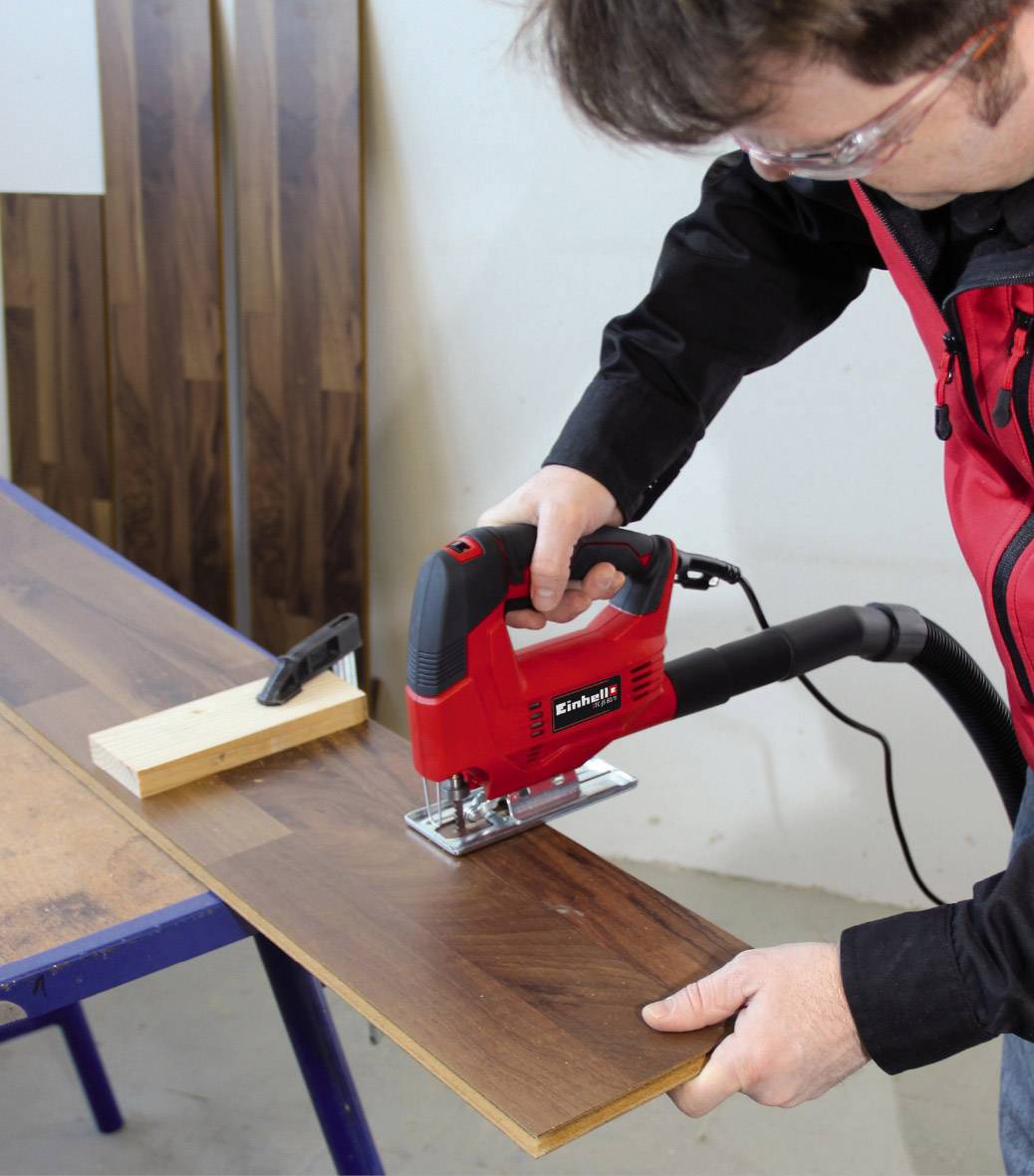 Une personne coupe une planche de bois avec une scie sauteuse sur une table. Les copeaux sont aspirés. D'autres planches sont disposées en arrière-plan.