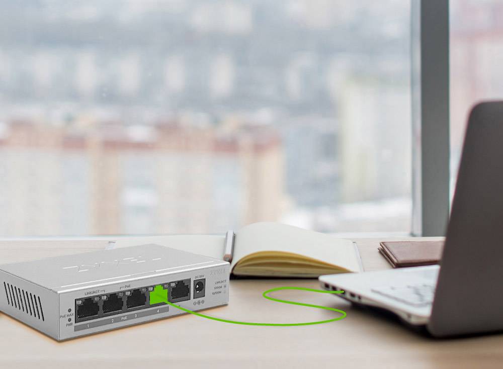 Un ordinateur portable est connecté à un commutateur réseau sur un bureau. En arrière-plan, on aperçoit un paysage urbain flou.