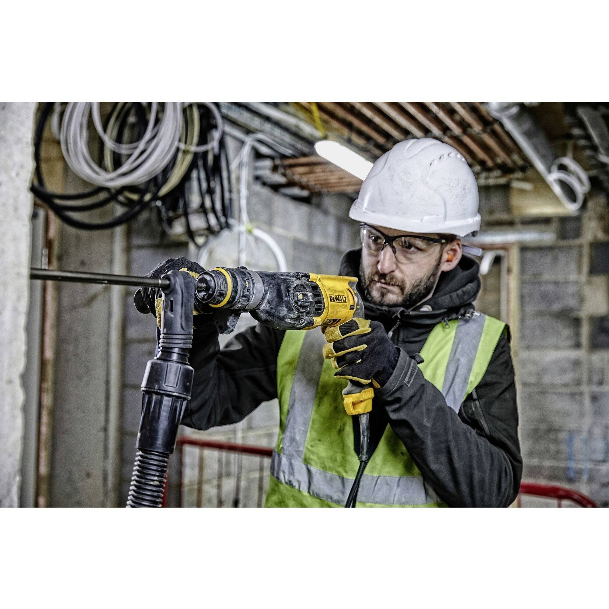 Un ouvrier portant des équipements de protection et un casque utilise une perceuse sur un chantier de construction. Des câbles et des tuyaux sont visibles en arrière-plan.