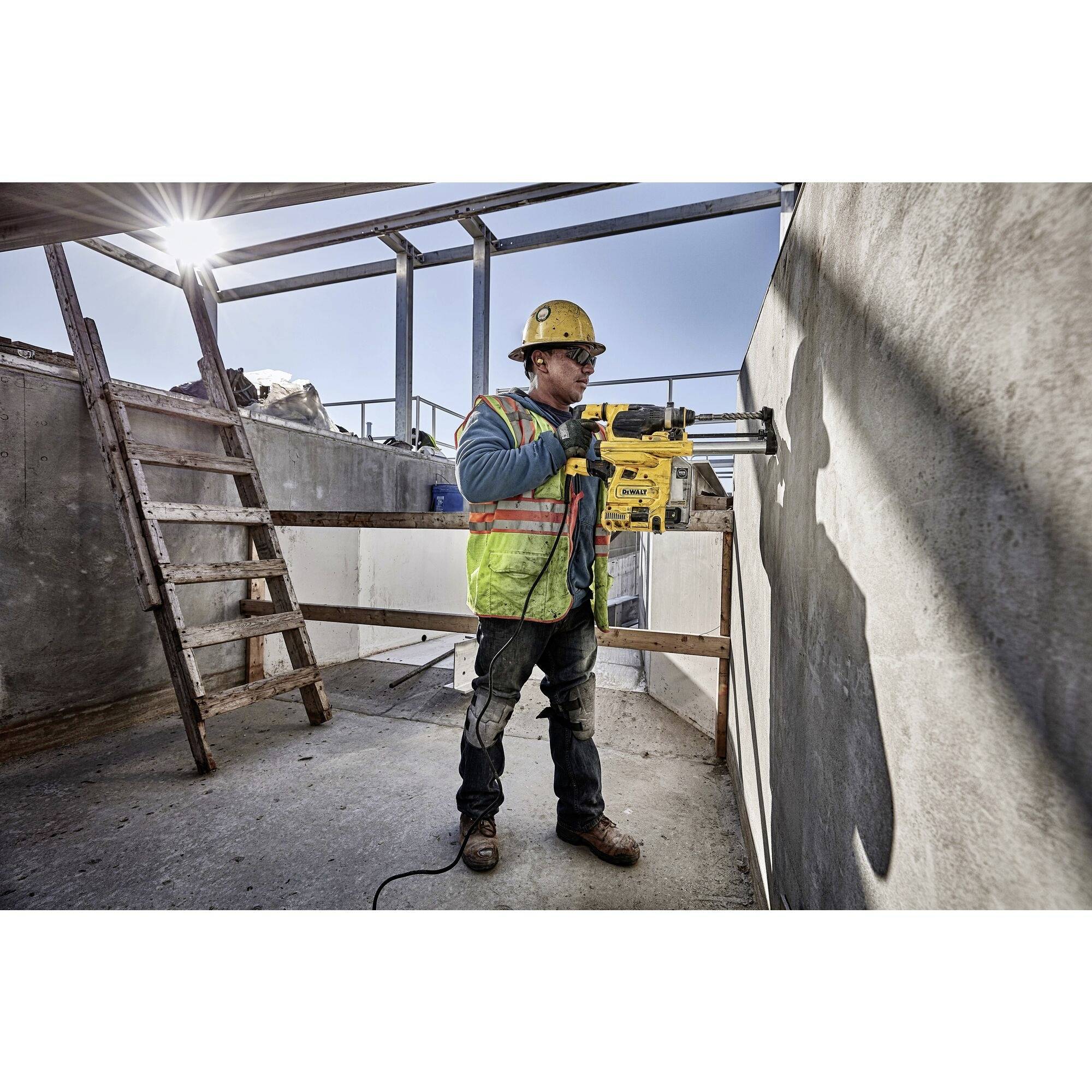 Un ouvrier portant un casque jaune et un gilet de sécurité utilise un outil électrique pour percer un mur en béton ; la lumière du soleil tombe de la gauche.