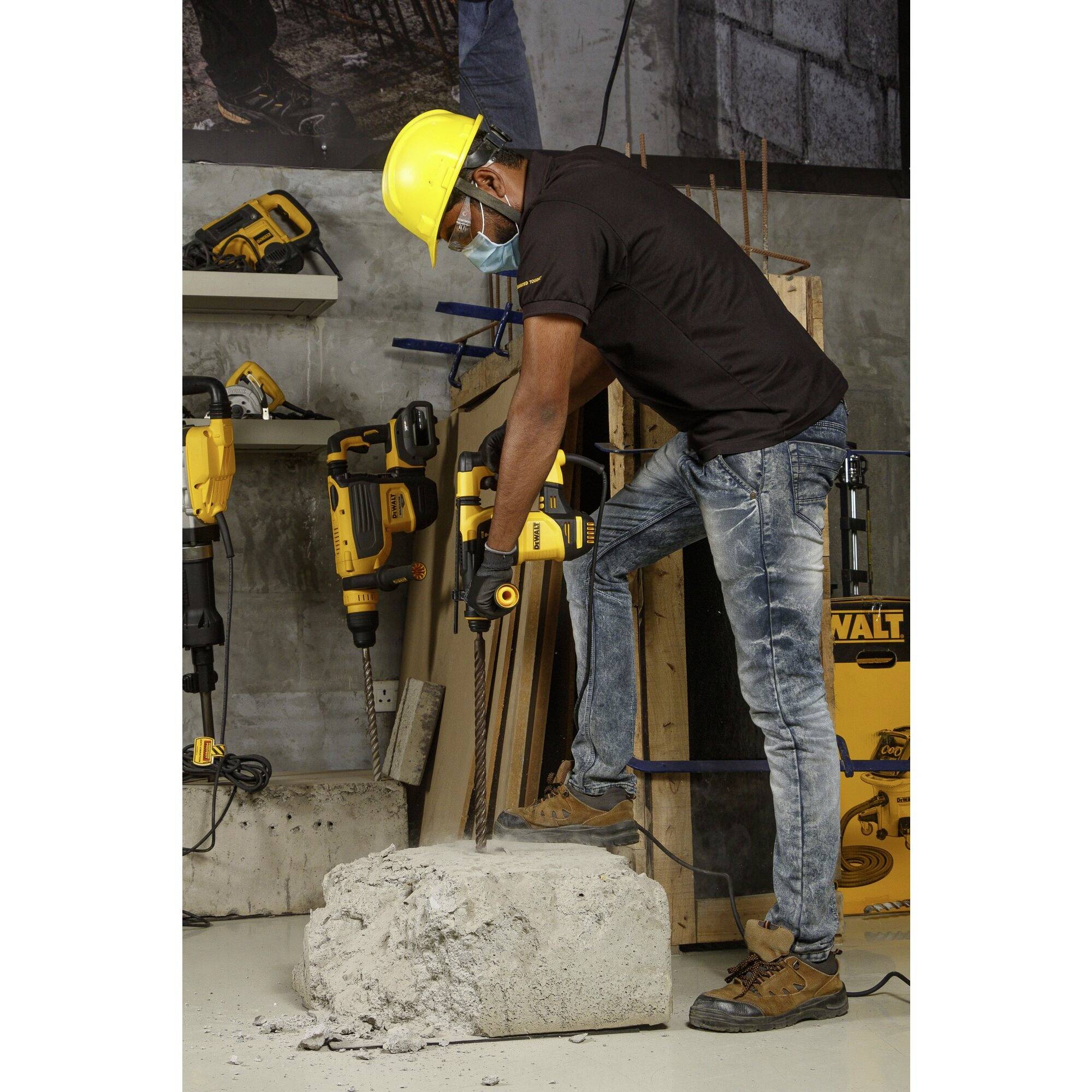 Un ouvrier portant un casque et un masque utilise une perceuse et travaille une pièce en béton dans un environnement d'atelier.