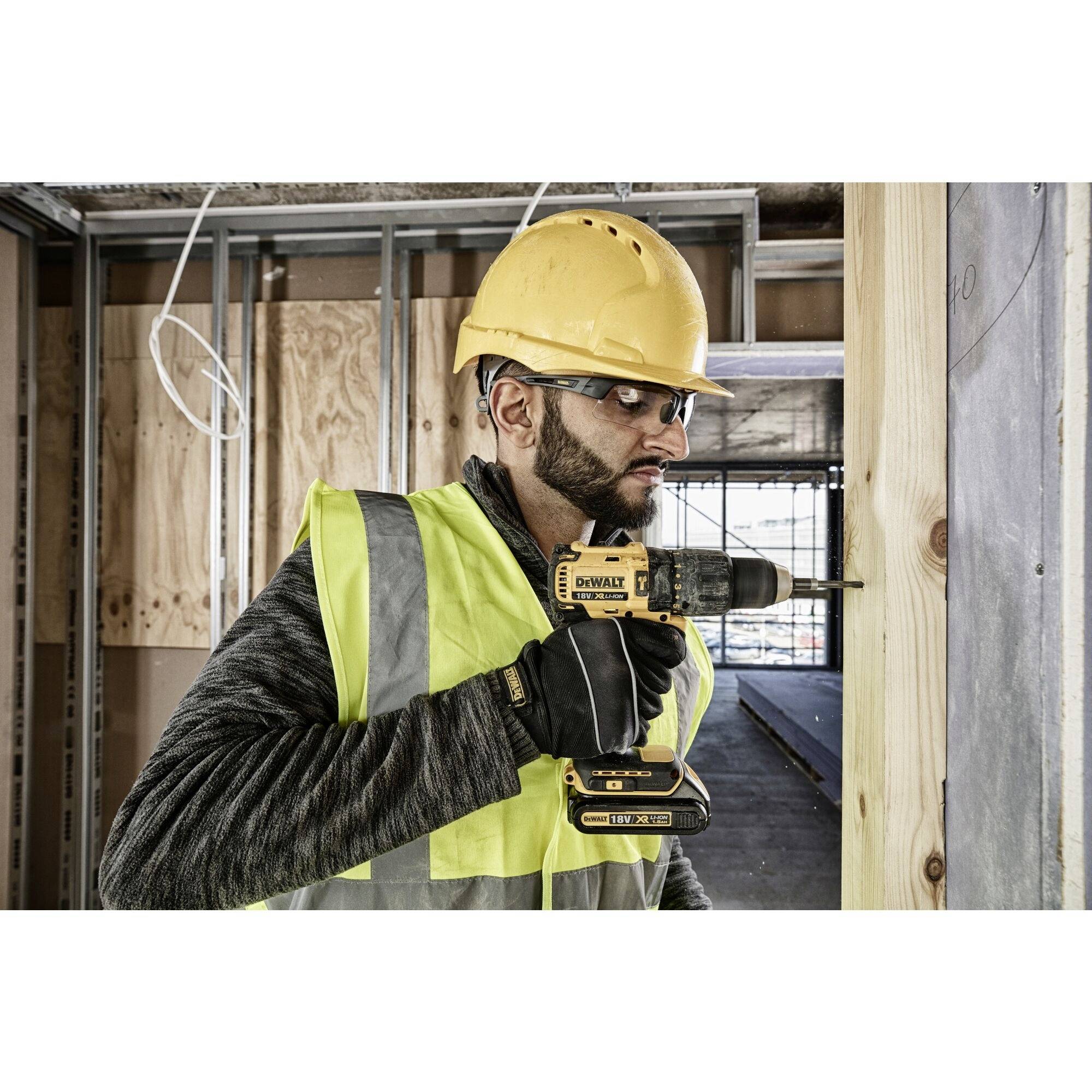 Un ouvrier portant un casque de chantier et un gilet de sécurité utilise une perceuse sur un poteau en bois sur un chantier de construction.