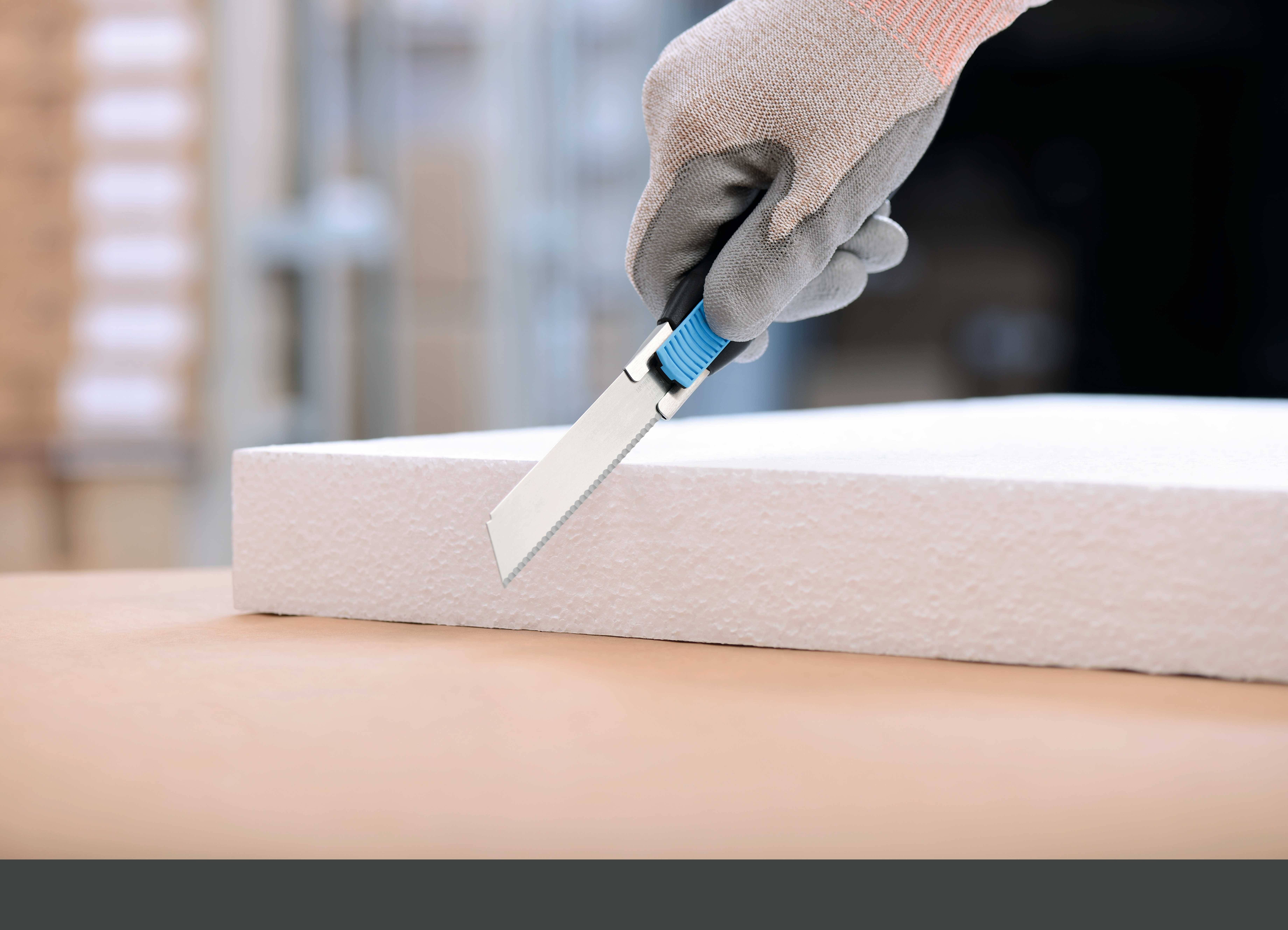 Une personne coupe une plaque de mousse avec un cutter. Le focus est mis sur la main gantée et le couteau.