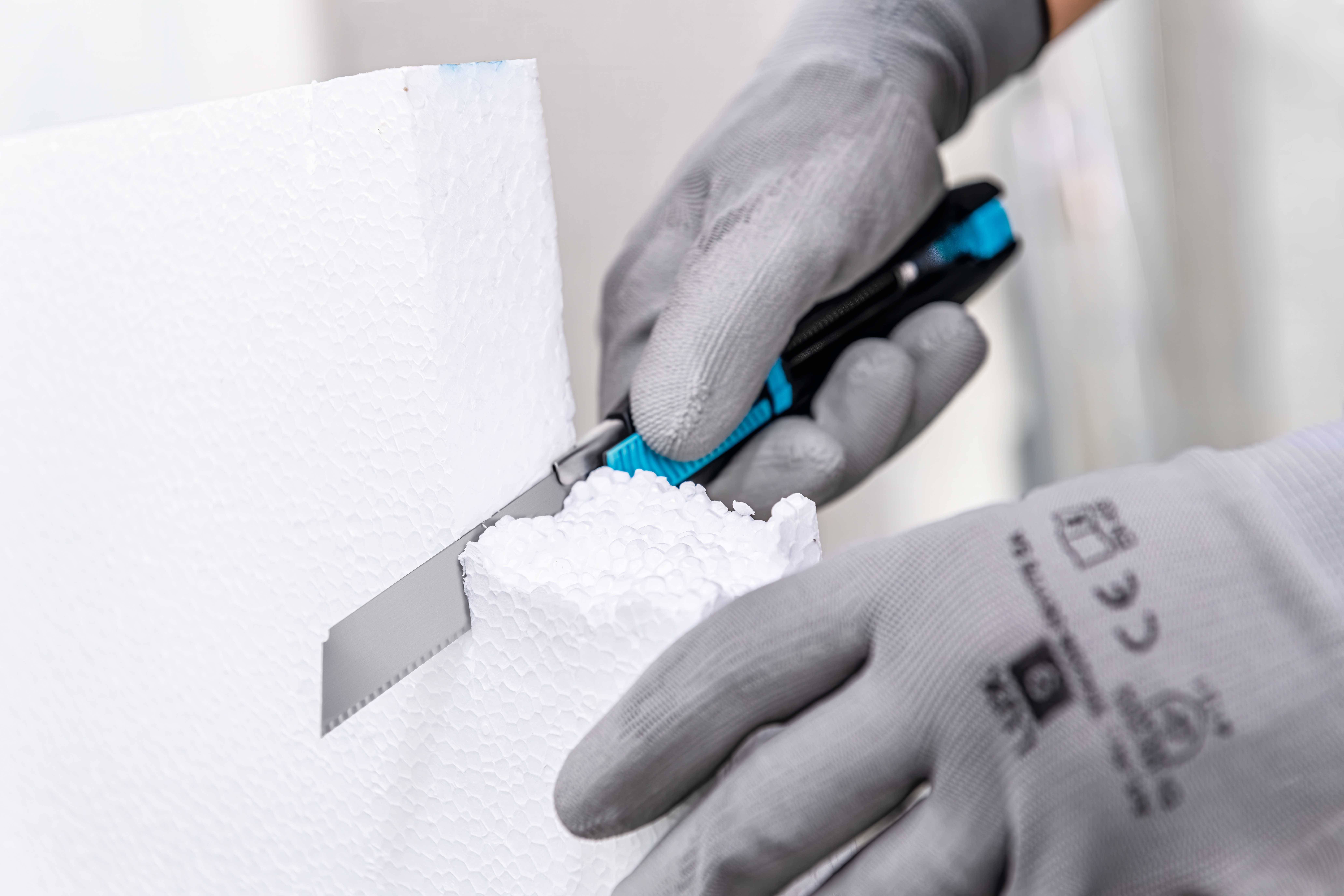Une personne coupe du polystyrène avec un petit couteau. Elle porte des gants gris pour protéger ses mains.