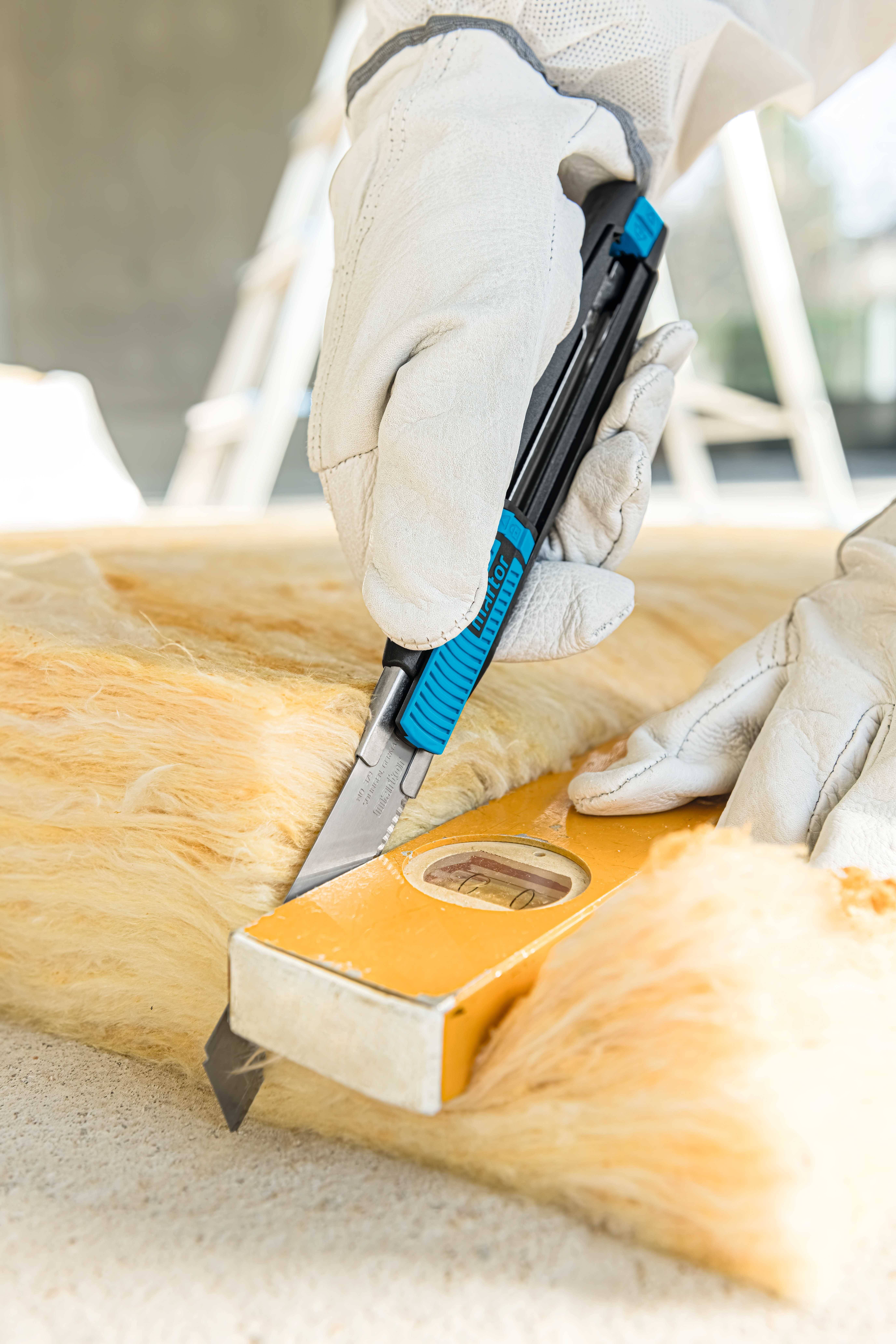 Une personne porte des gants de travail blancs et coupe un morceau de tapis isolant jaune à l'aide d'un cutter sur un niveau à bulle.