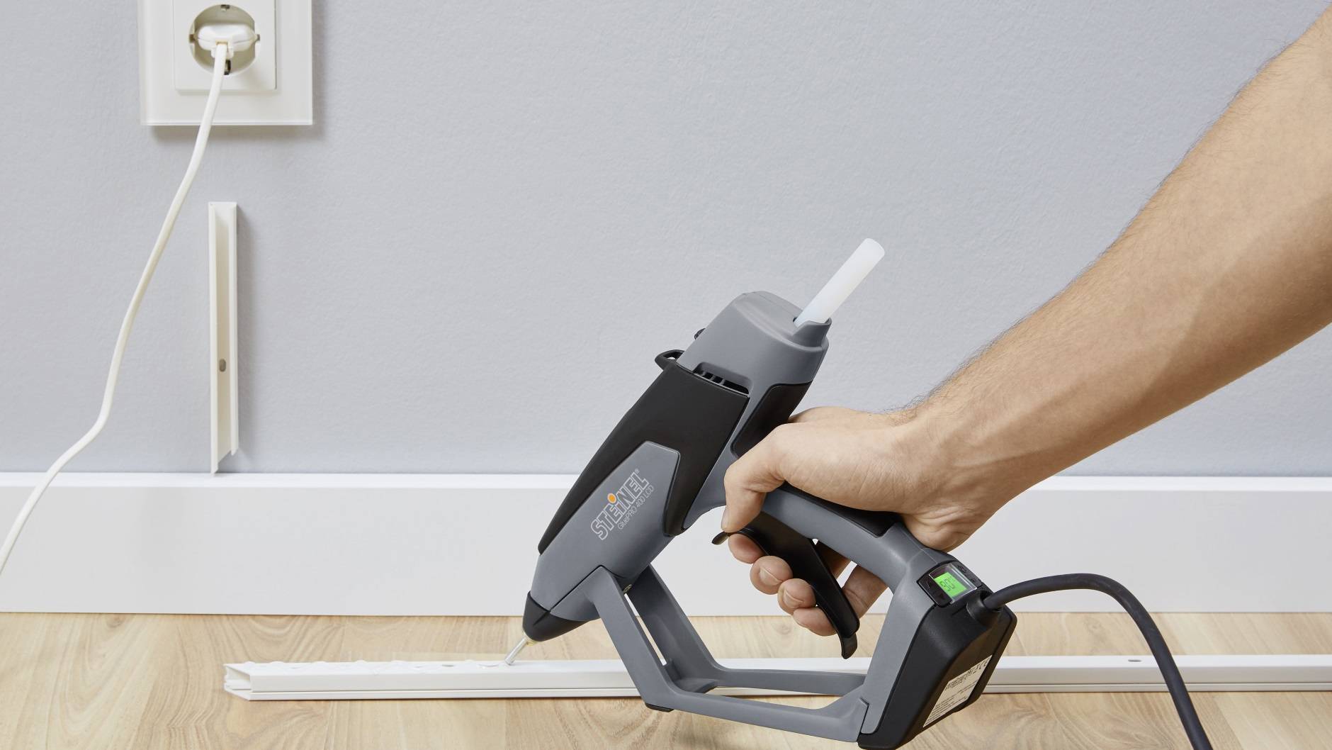 Une personne tient un pistolet à colle chaude et fixe un cache-câble le long d'une plinthe. Une prise électrique est visible en arrière-plan.