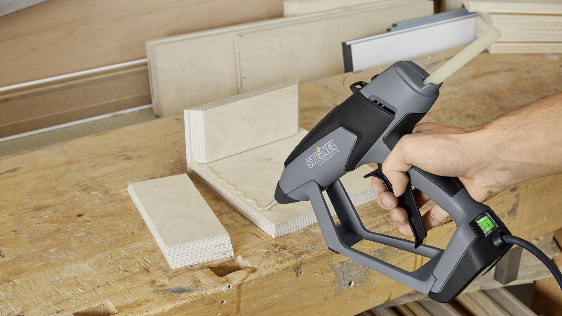 Une main tient un pistolet à colle chaude au-dessus d'une table en bois, prête à être utilisée sur des morceaux de bois empilés en arrière-plan.