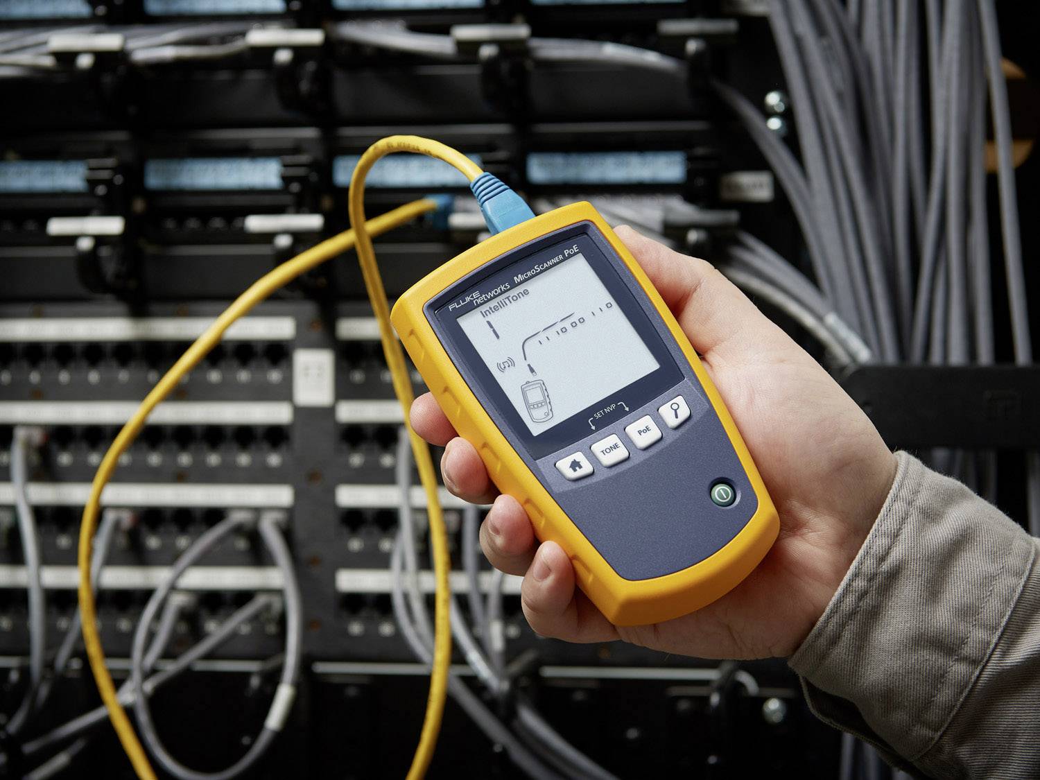 Une personne tient un appareil de test de câbles devant un coffret de distribution réseau. L'écran affiche une situation de test de connexion de câble.