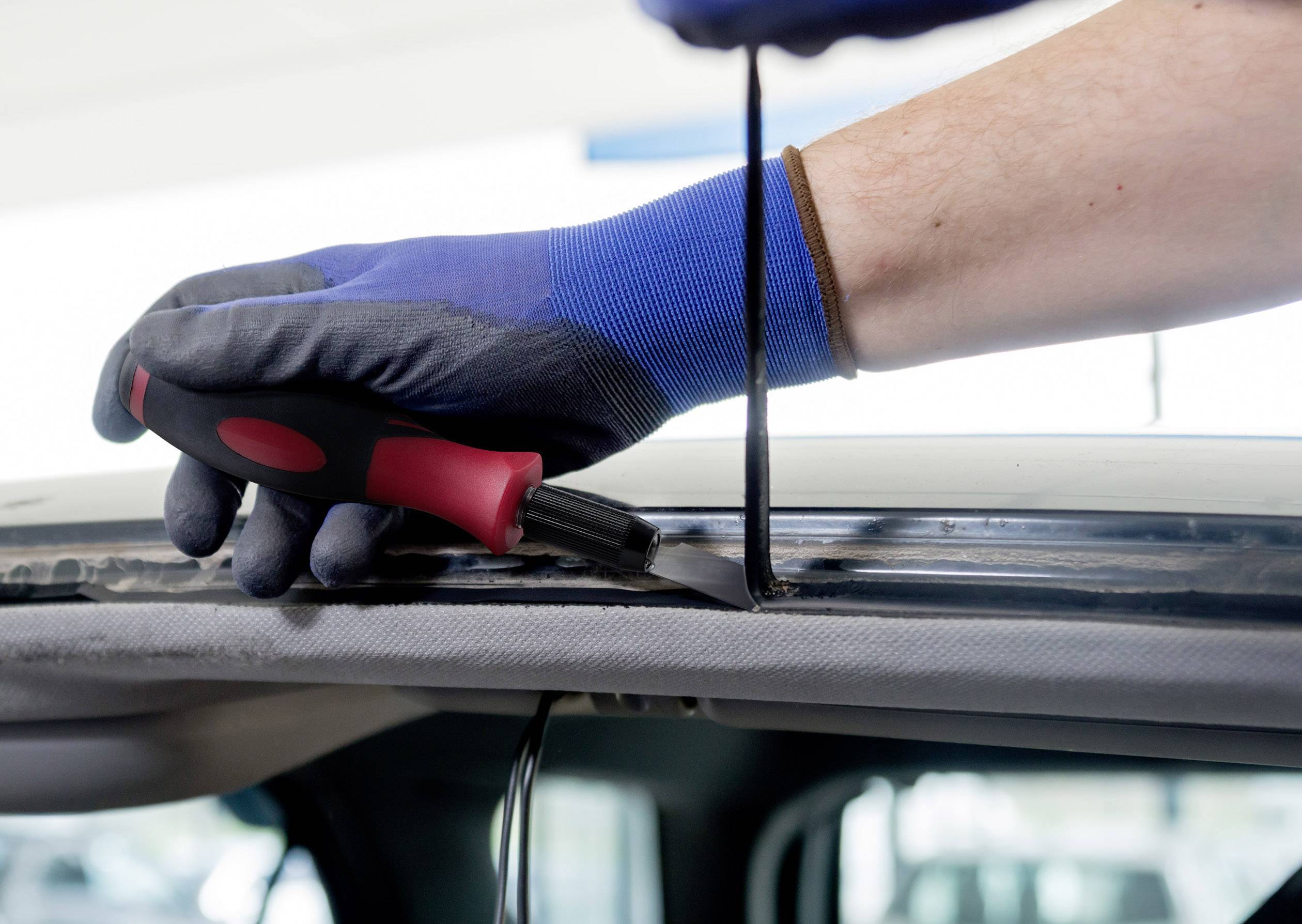 Quelqu'un utilise un couteau pour retirer le joint d'étanchéité d'une vitre de voiture. La personne porte des gants de travail bleus.