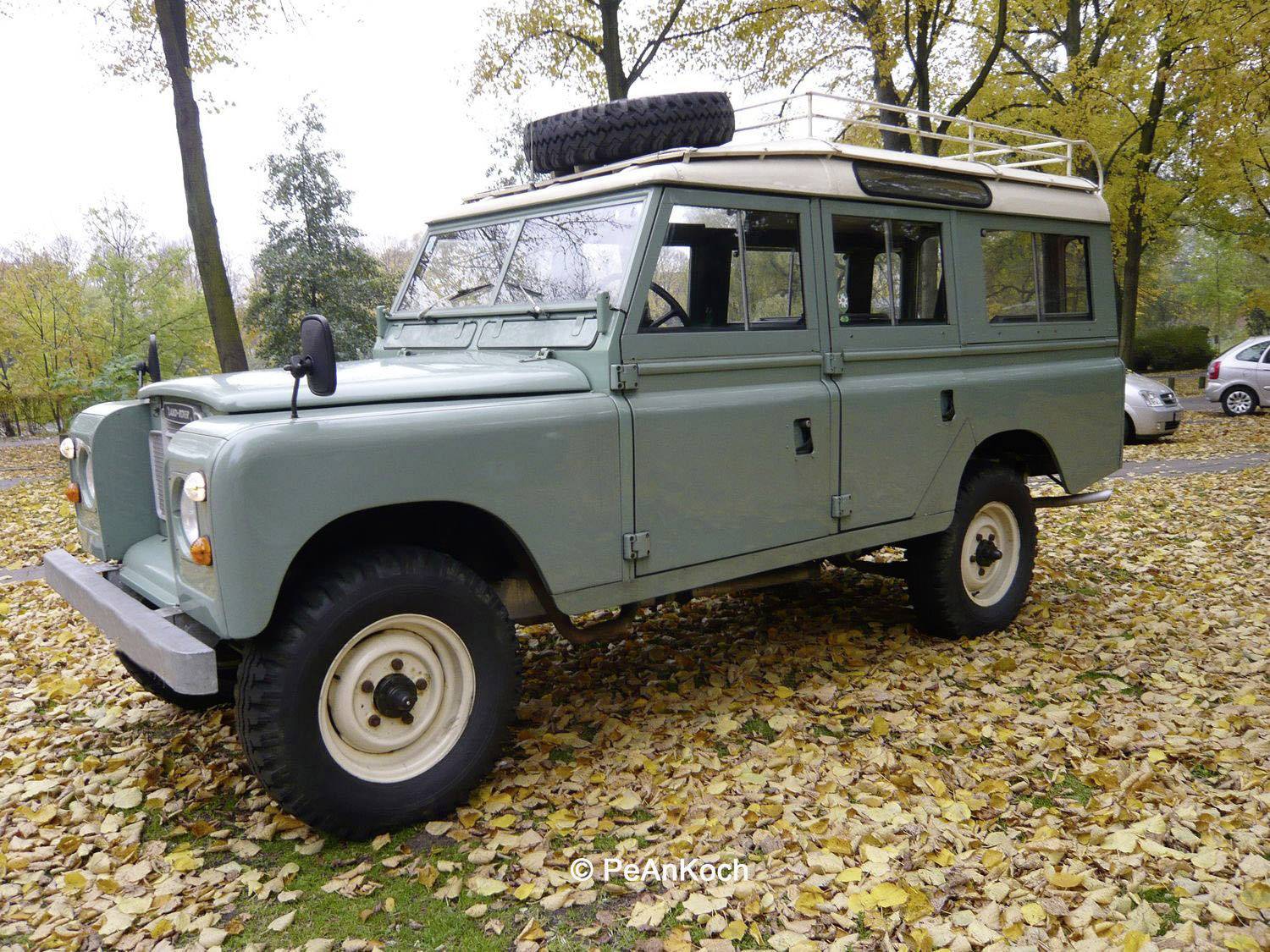 Un véhicule tout-terrain classique, vert clair, est stationné sur une prairie couverte de feuilles dans un parc.