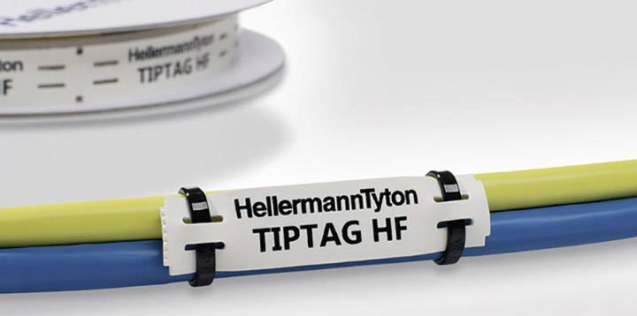 Une étiquette de câble blanche portant l'inscription 'HellermannTyton TIPTAG HF' est fixée sur des câbles jaunes et bleus. En arrière-plan, on aperçoit un bobine.