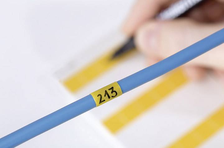 Une main tient un stylo au-dessus d'une feuille portant des diagrammes jaunes. Au premier plan, un câble bleu portant le numéro '213'.