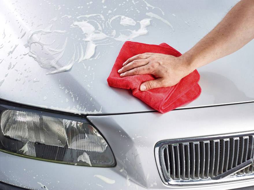 Une main tenant un chiffon rouge polit le capot d'une voiture argentée, recouverte d'eau savonneuse.