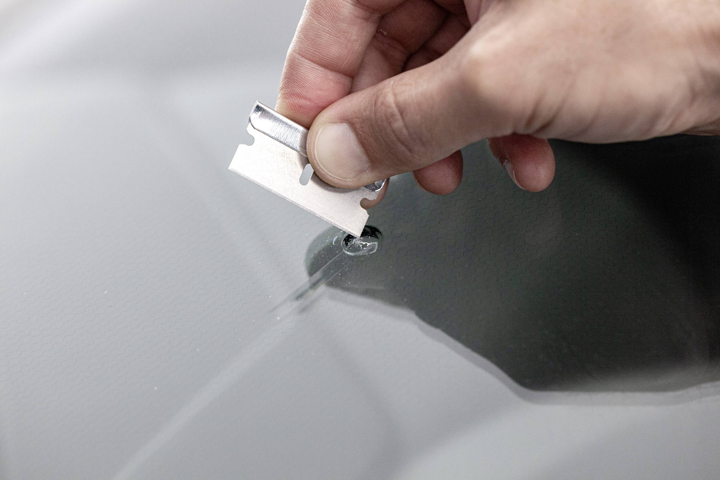 Une main tient une lame de rasoir au-dessus d'un éclat de verre sur un pare-brise de voiture, prête à enlever le fragment.