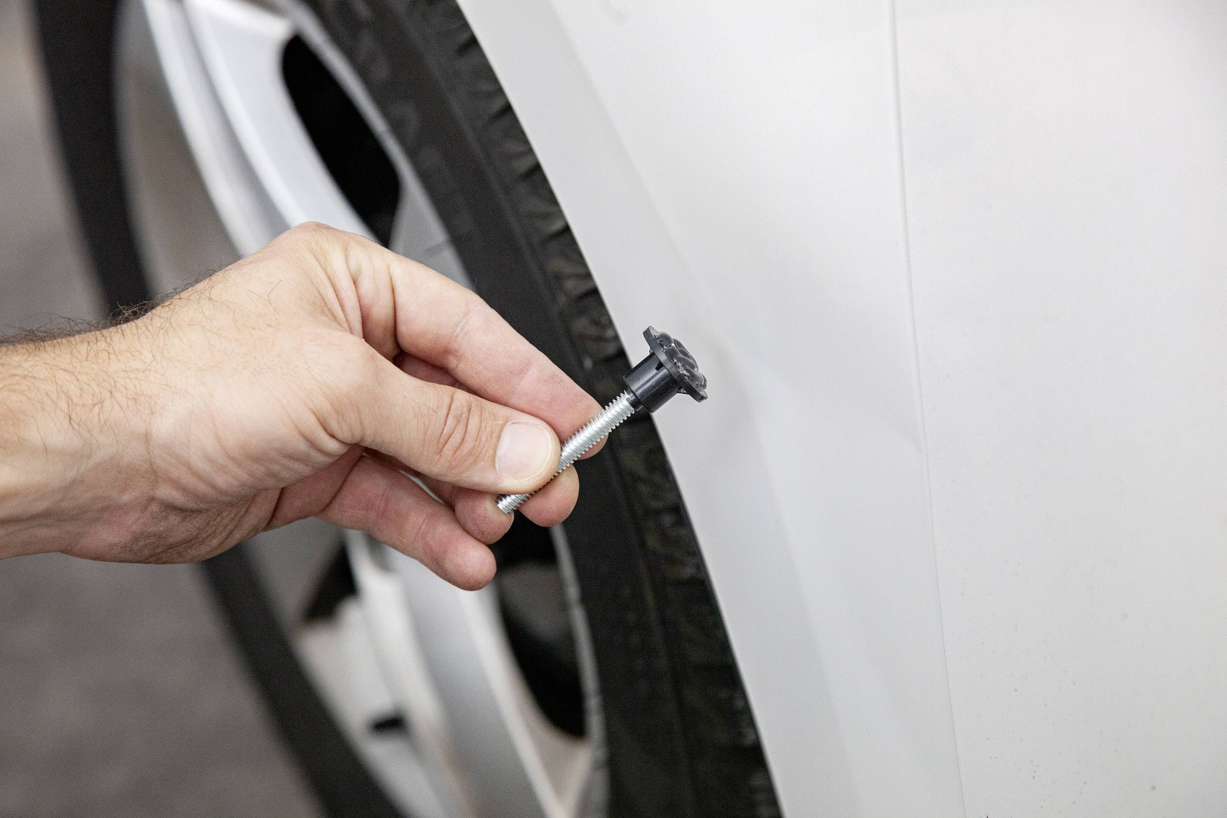 Une main tient une cheville d'ancrage près d'un pneu de voiture, alors qu'une rayure est visible sur la carrosserie.