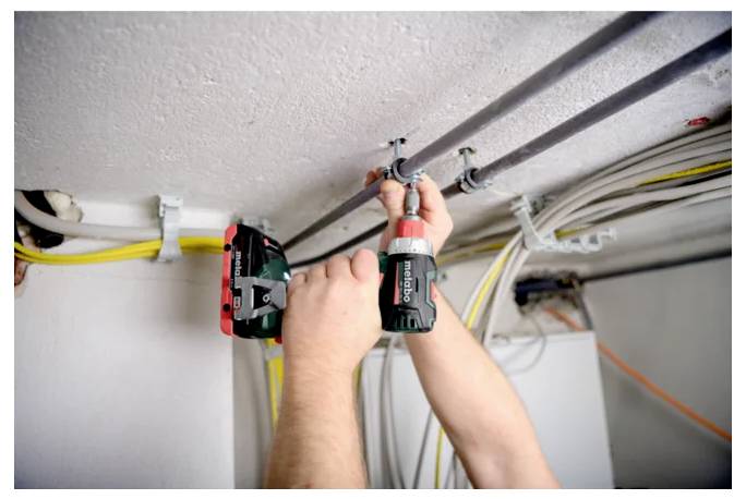 Électricien utilisant une perceuse pour fixer une gaine au plafond, avec des câbles exposés dans une pièce inachevée.