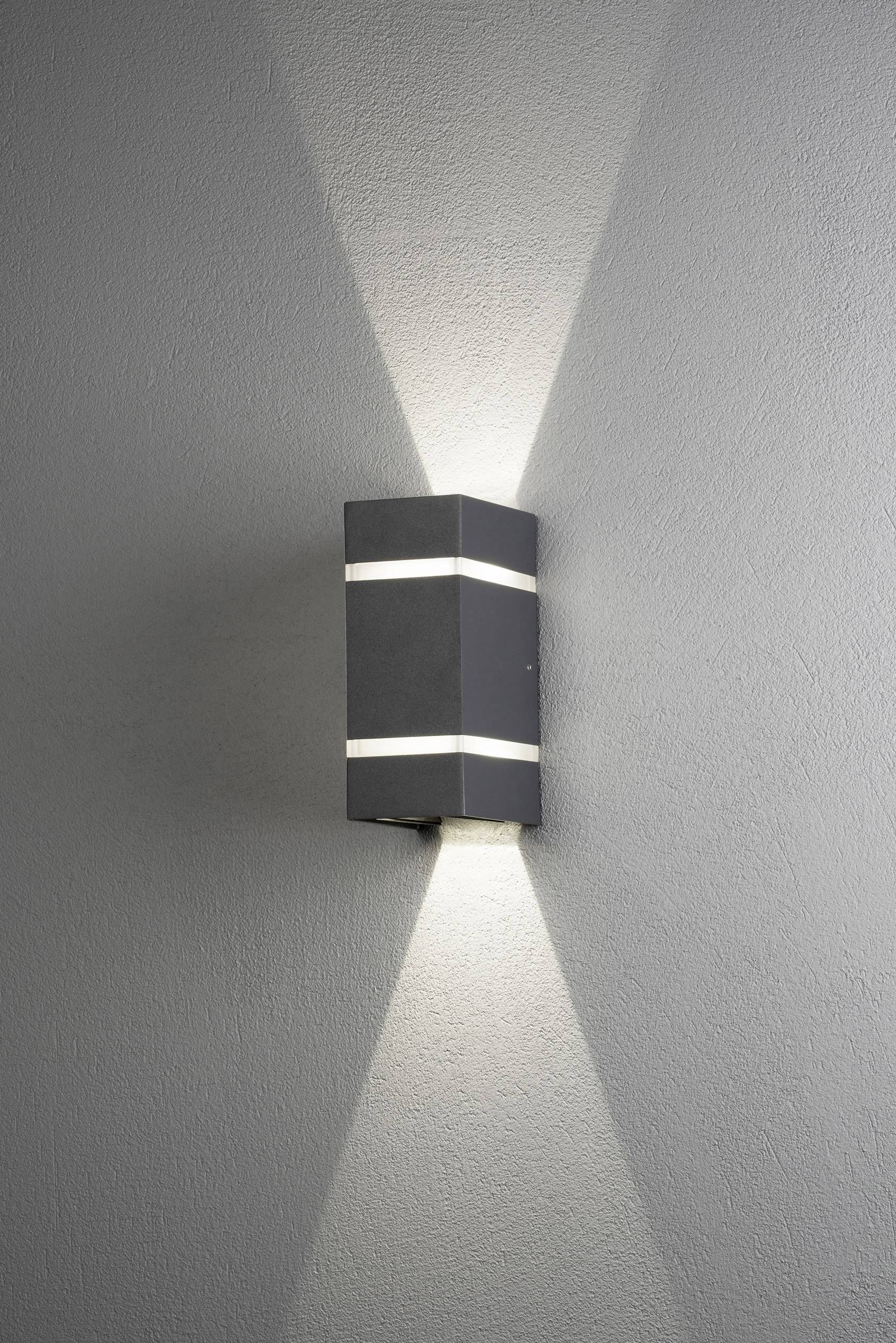 Un applique murale rectangulaire noir éclaire vers le haut et vers le bas, créant un motif lumineux symétrique sur le mur gris.