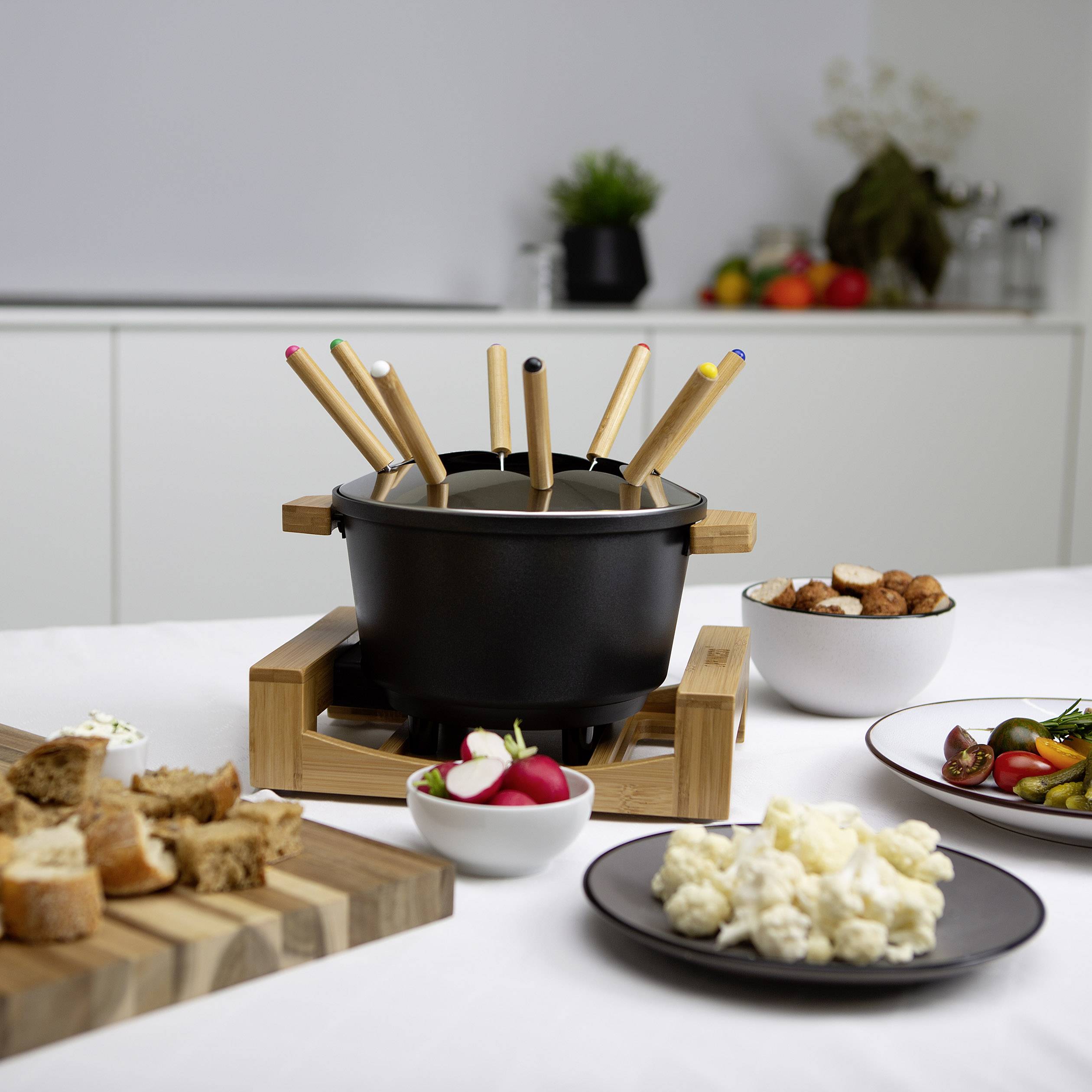 Un set à fondue avec des casseroles et des fourchettes en bois sur une table. En arrière-plan, on aperçoit des assiettes de légumes, de pain et de sauces.