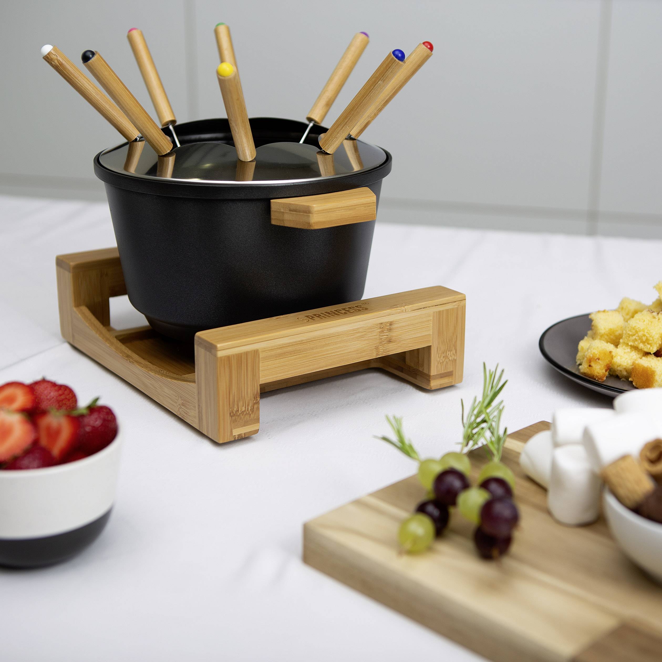 Un caquelon de fondue avec plusieurs fourchettes à fondue est posé sur une table. À côté se trouvent des coupes de fruits et de petites collations, prêts pour la dégustation de fondue.